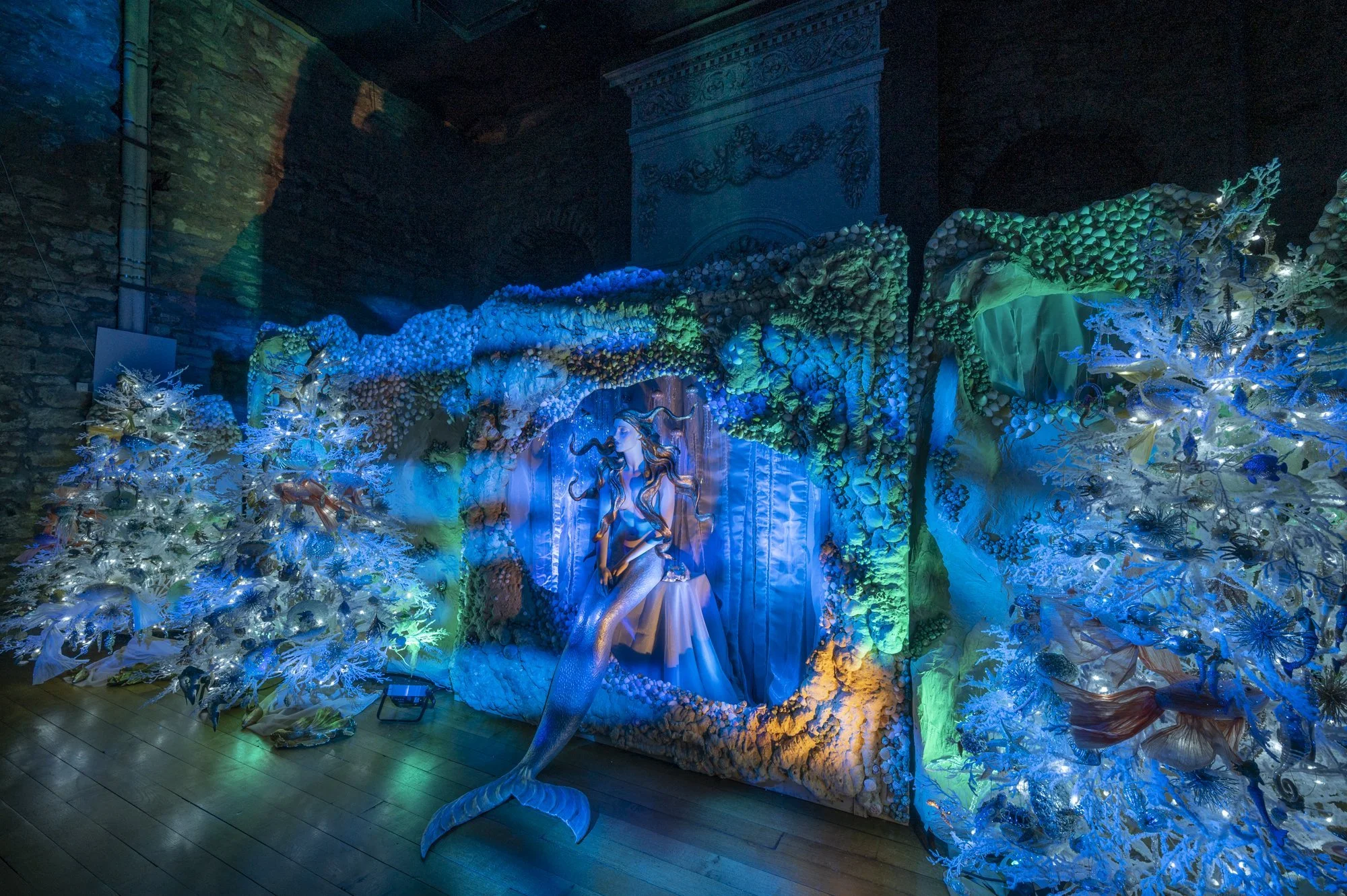Colorful art installation featuring a mermaid sculpture surrounded by coral and seaweed, illuminated with blue and green lights, in a dimly lit room with brick and ornate decorative walls.