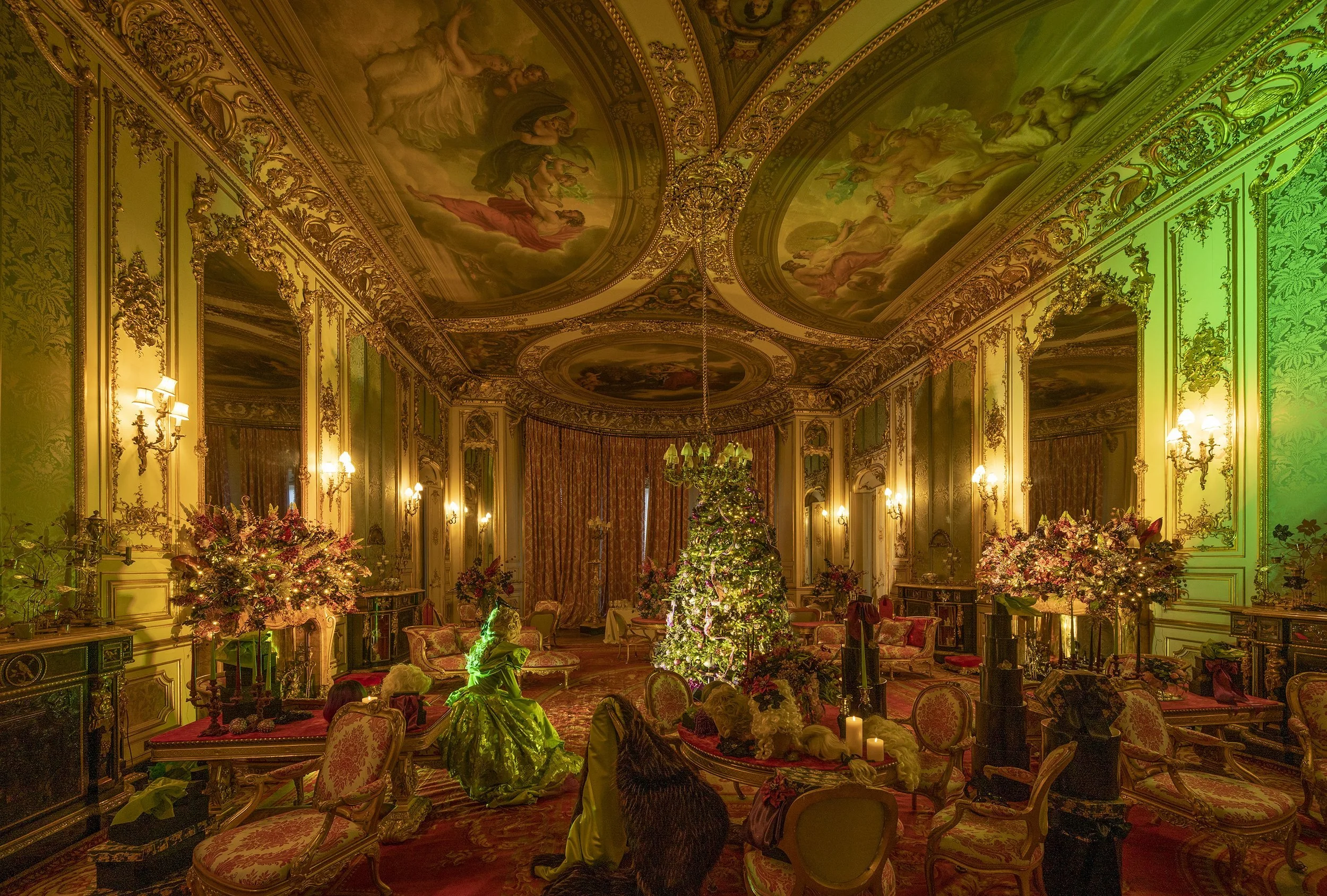 Luxurious room decorated for Christmas with a decorated tree, floral arrangements, Christmas figures, and ornate gold-accented walls and ceiling murals.