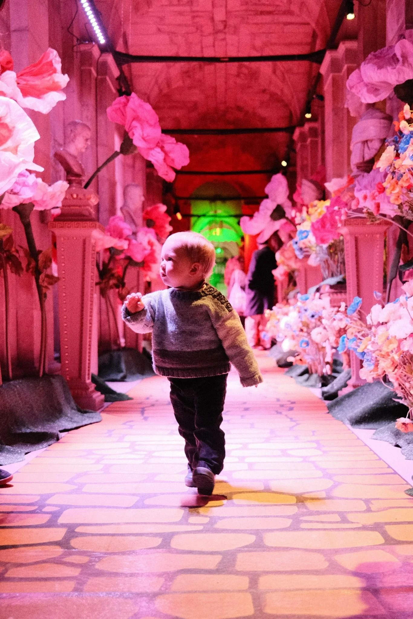 A young boy with blonde hair walking down a colorful, illuminated hallway decorated with pink flowers and floral arrangements on both sides. The hallway has pink and purple lighting with brick and stone decor and a high, arched ceiling with green and