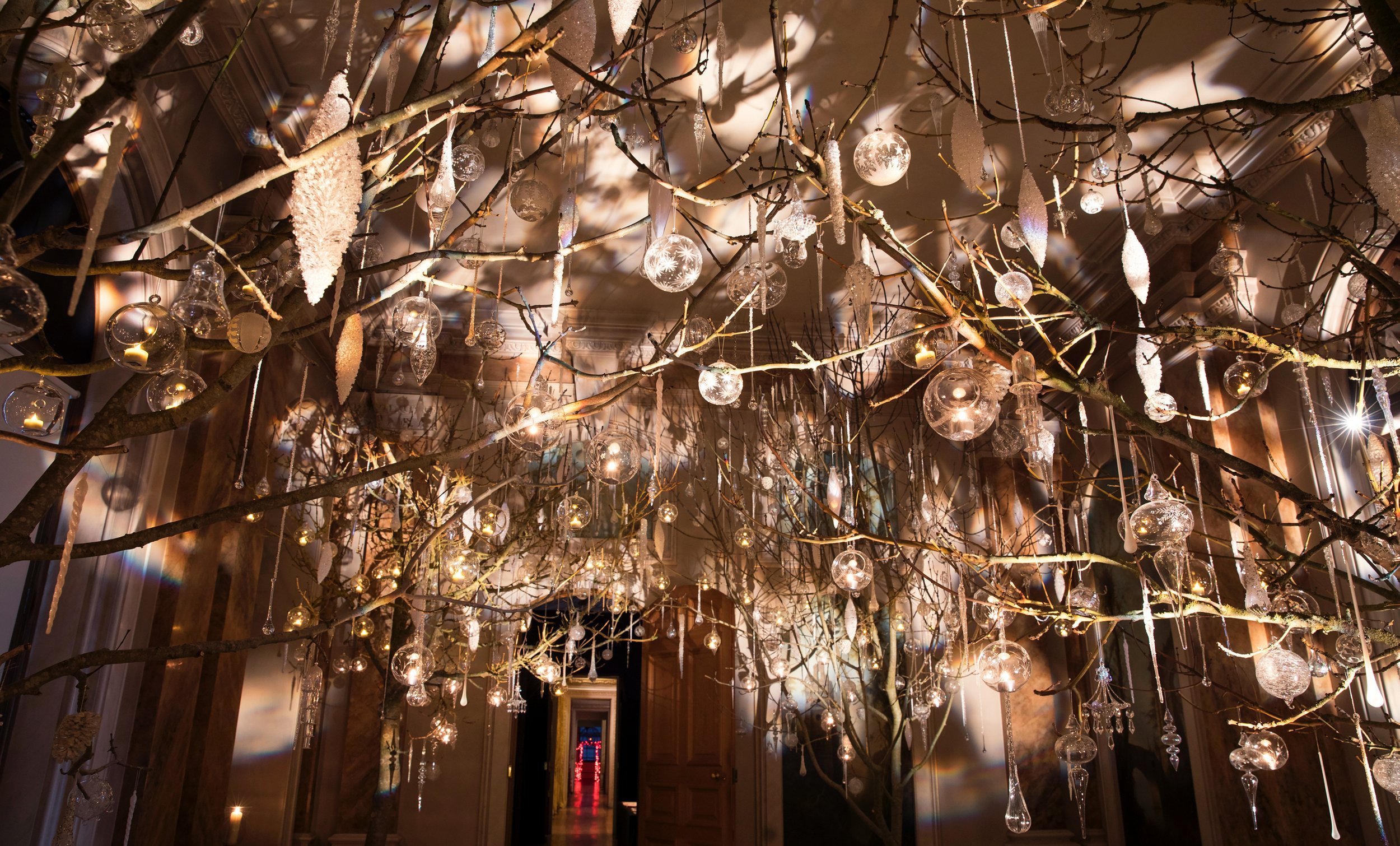 An elaborate indoor ceiling decoration featuring bare tree branches adorned with clear glass ornaments, icicle-shaped decorations, and hanging candles, illuminated with warm lighting.