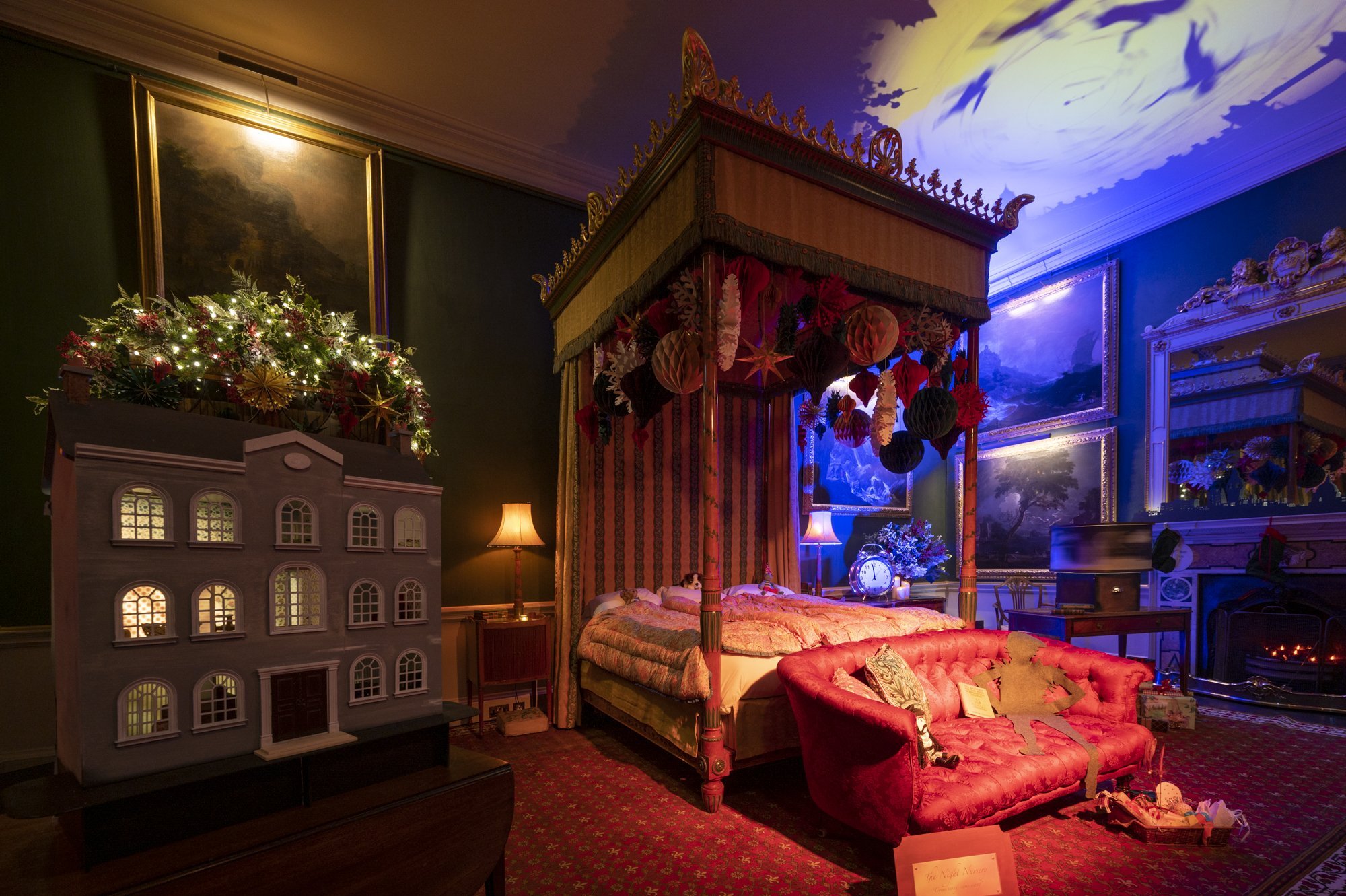 A bedroom decorated for Christmas with a four-poster bed, red velvet furniture, a chandelier, and Christmas ornaments hanging from the ceiling, along with paintings and holiday decorations.