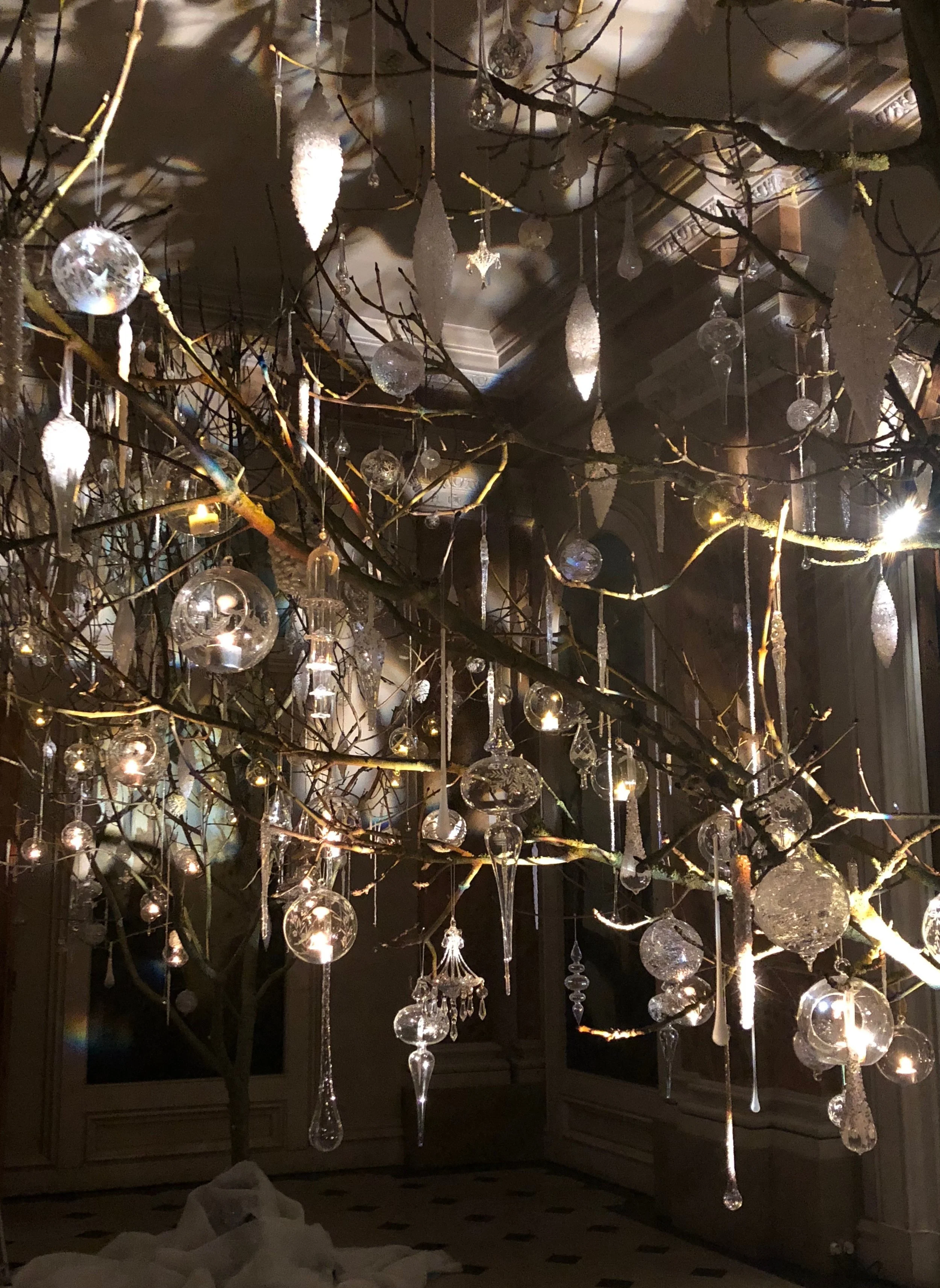 A chandelier made of branches with numerous hanging glass ornaments and candles inside a dark room.