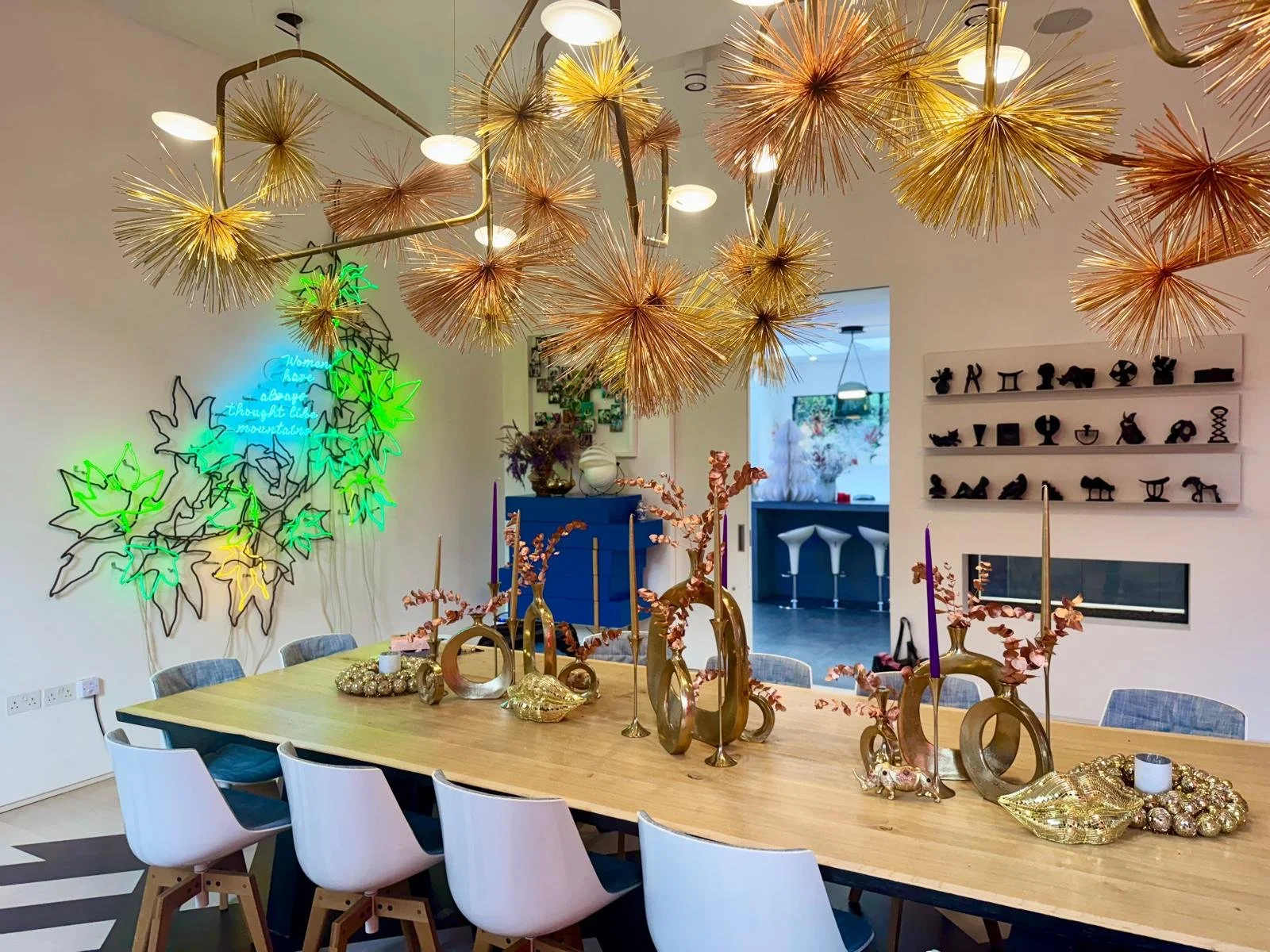 A dining room decorated with a large wooden table, gold and pink vases with pink branches, gold candle holders, and modern gold decor. The ceiling has a large, artistic gold light fixture. On the wall there is a neon sign with green, blue, and yellow