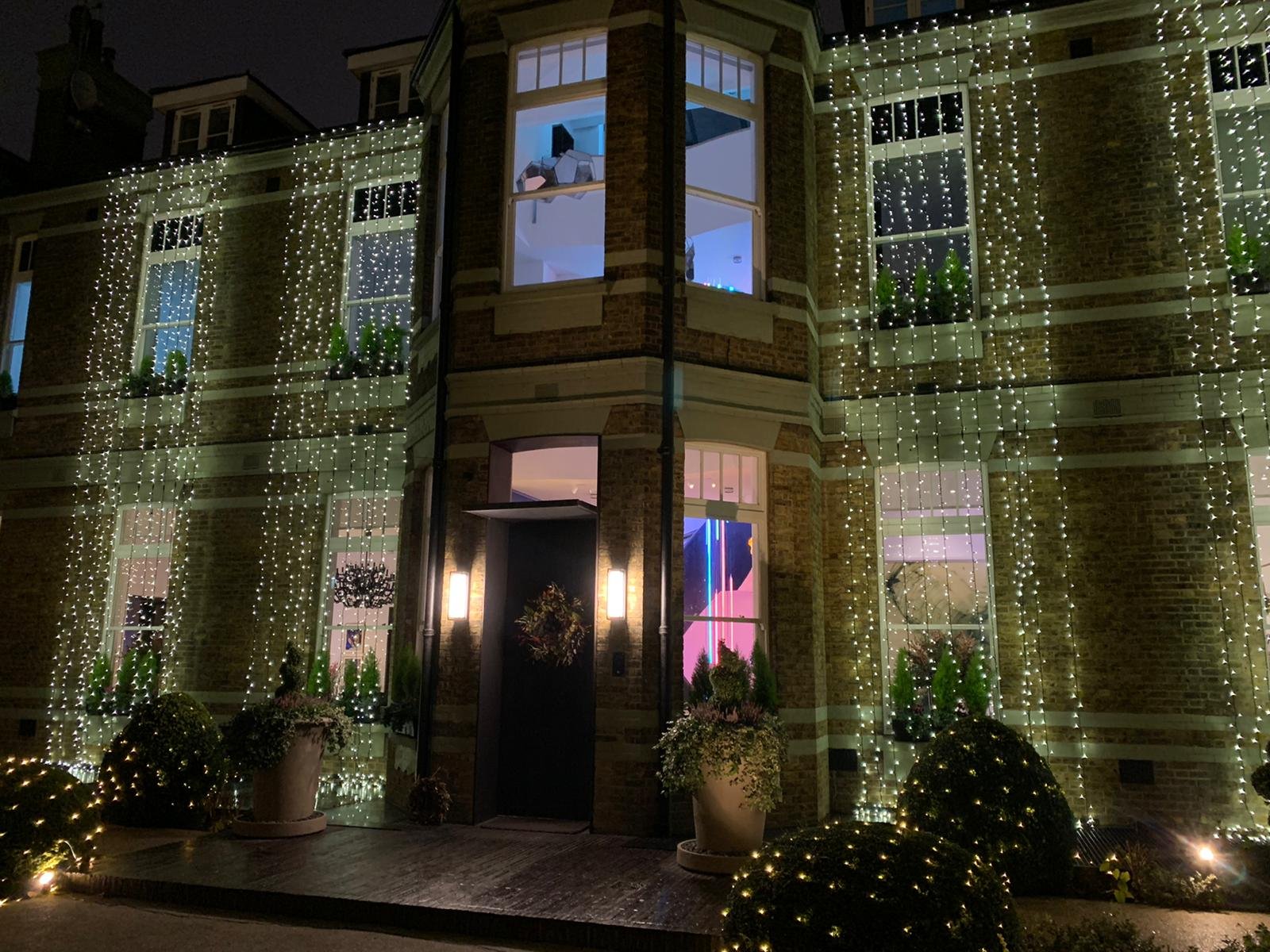 A multi-story house decorated with string lights around the windows and entrance at night. Potted plants and shrubs are visible in the front yard, illuminated by additional outdoor lighting.
