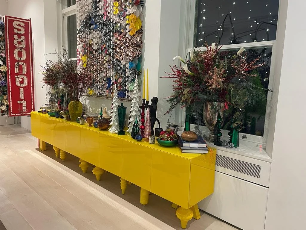 A bright yellow sideboard decorated with vases, books, and Christmas ornaments. Two large floral arrangements are on top, one near the window and one beside a colorful abstract wall hanging. A red banner with white lettering is partially visible on t