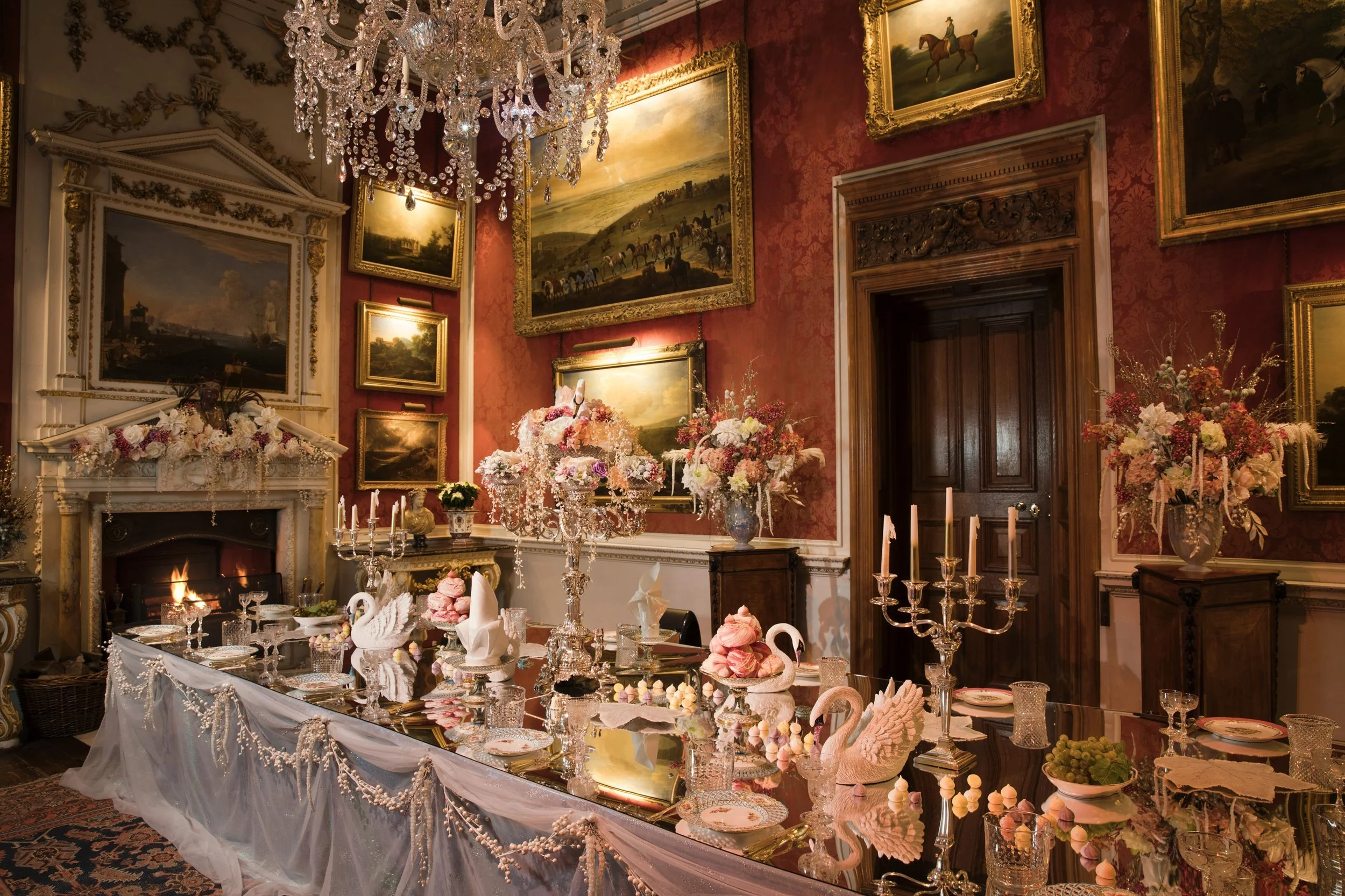 An elegantly decorated dining room with numerous framed landscape paintings on red wallpapered walls, a chandelier hanging from the ceiling, a fireplace with floral arrangements, and a table set for a celebration with pink and white decorations, cand