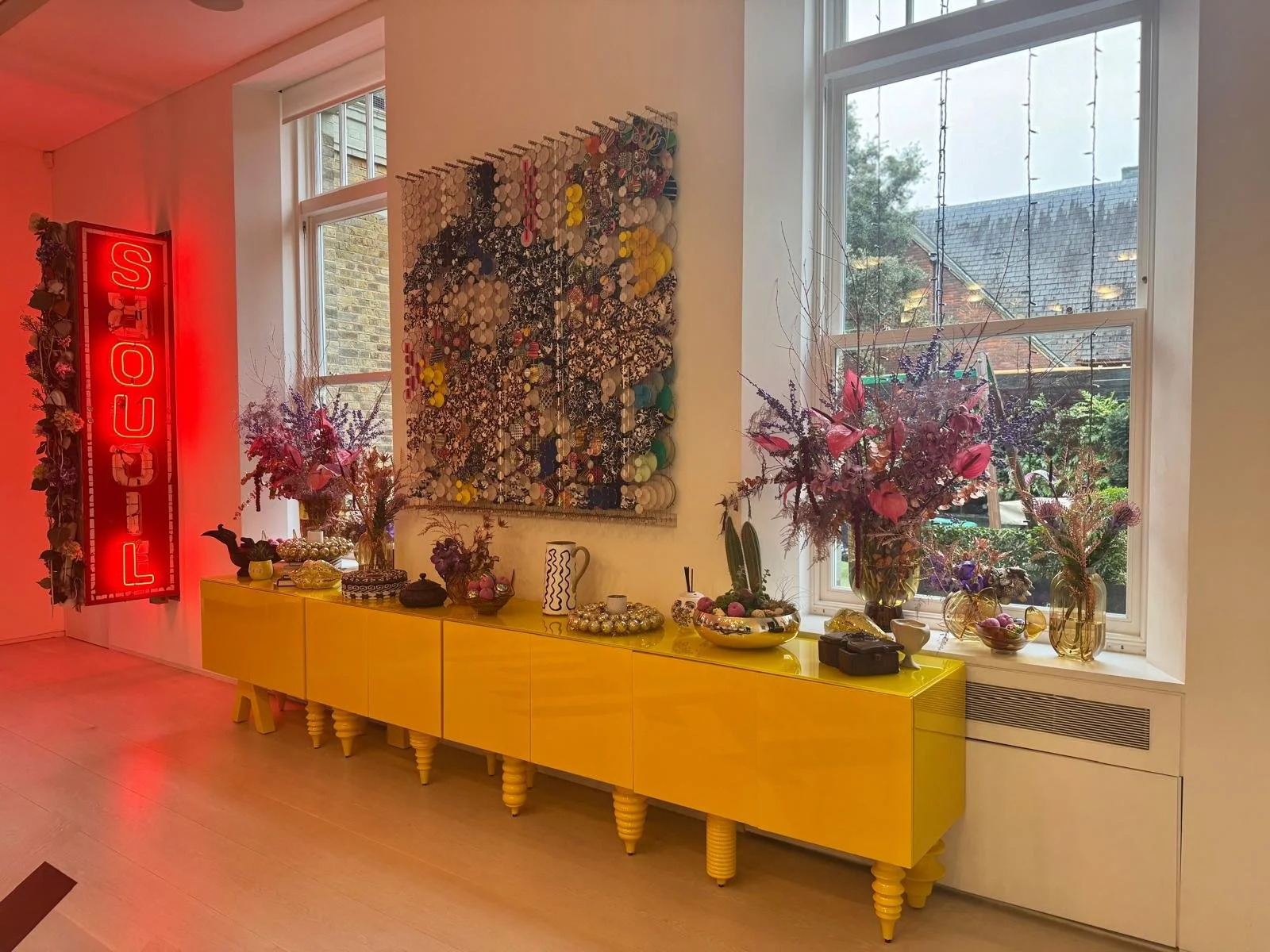 Decorative interior with a yellow sideboard, floral arrangements, a large window, and a neon red sign that says 'LOUIS' on the wall.