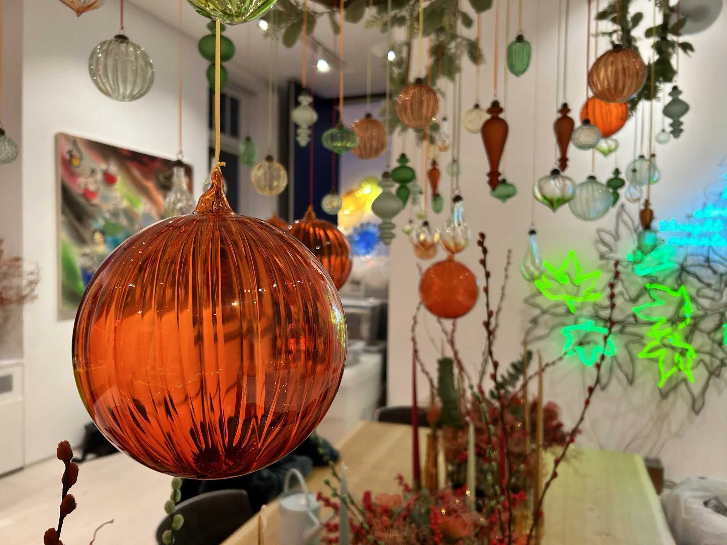 Close-up of colorful glass ornaments hanging, with a blurred background of more ornaments and holiday decorations.