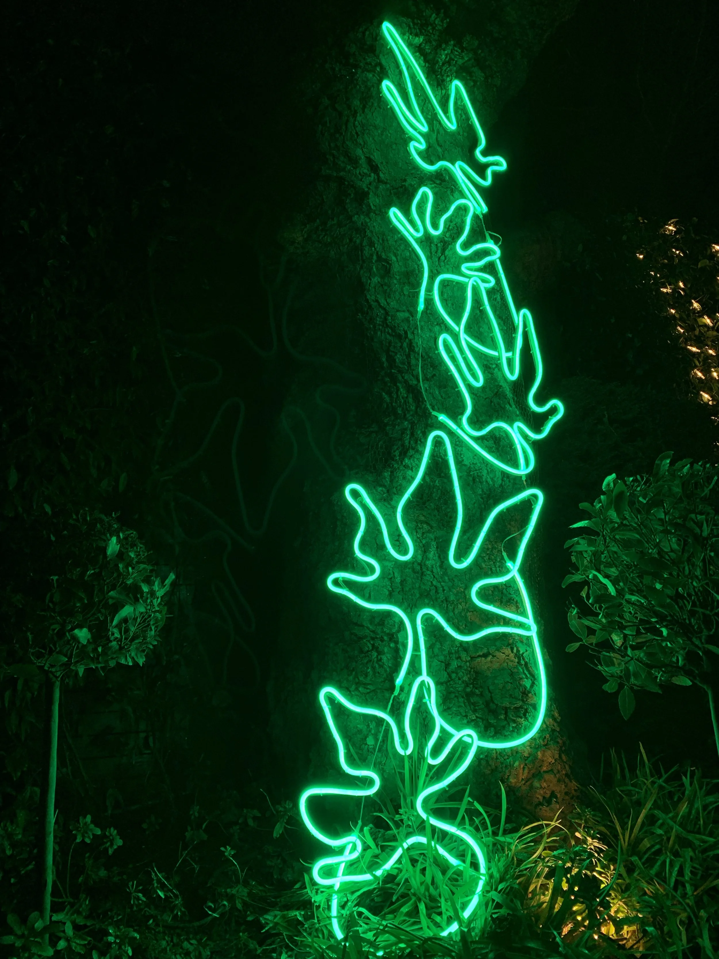 Green neon art installation of a bird with outstretched wings perched on a branch, mounted on a tree trunk, surrounded by plants at night.