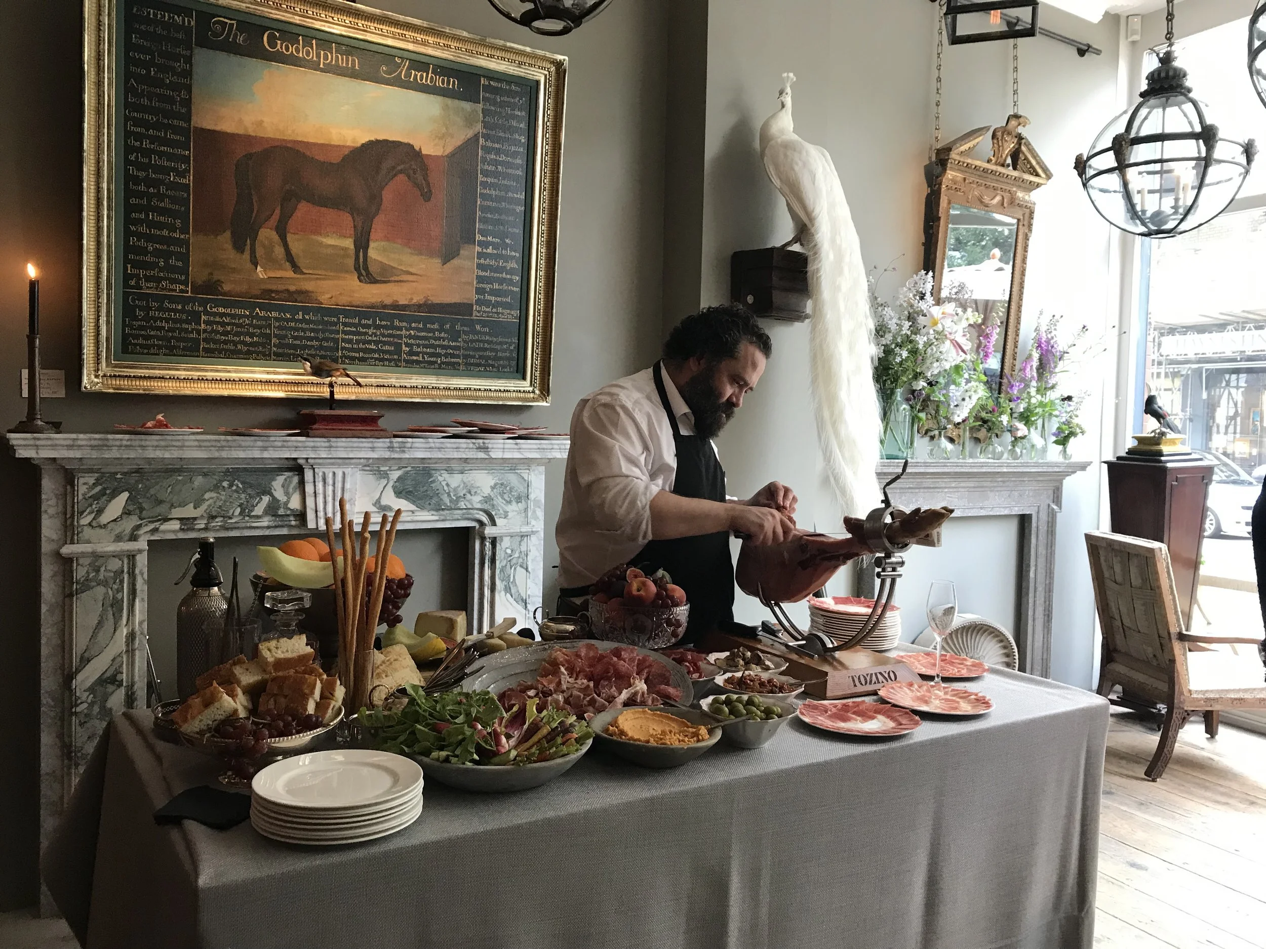 Buffet table with various appetizers and snacks, including sliced meats, cheeses, bread, fruit, and vegetables, set in a restaurant or café with a man preparing food in the background.