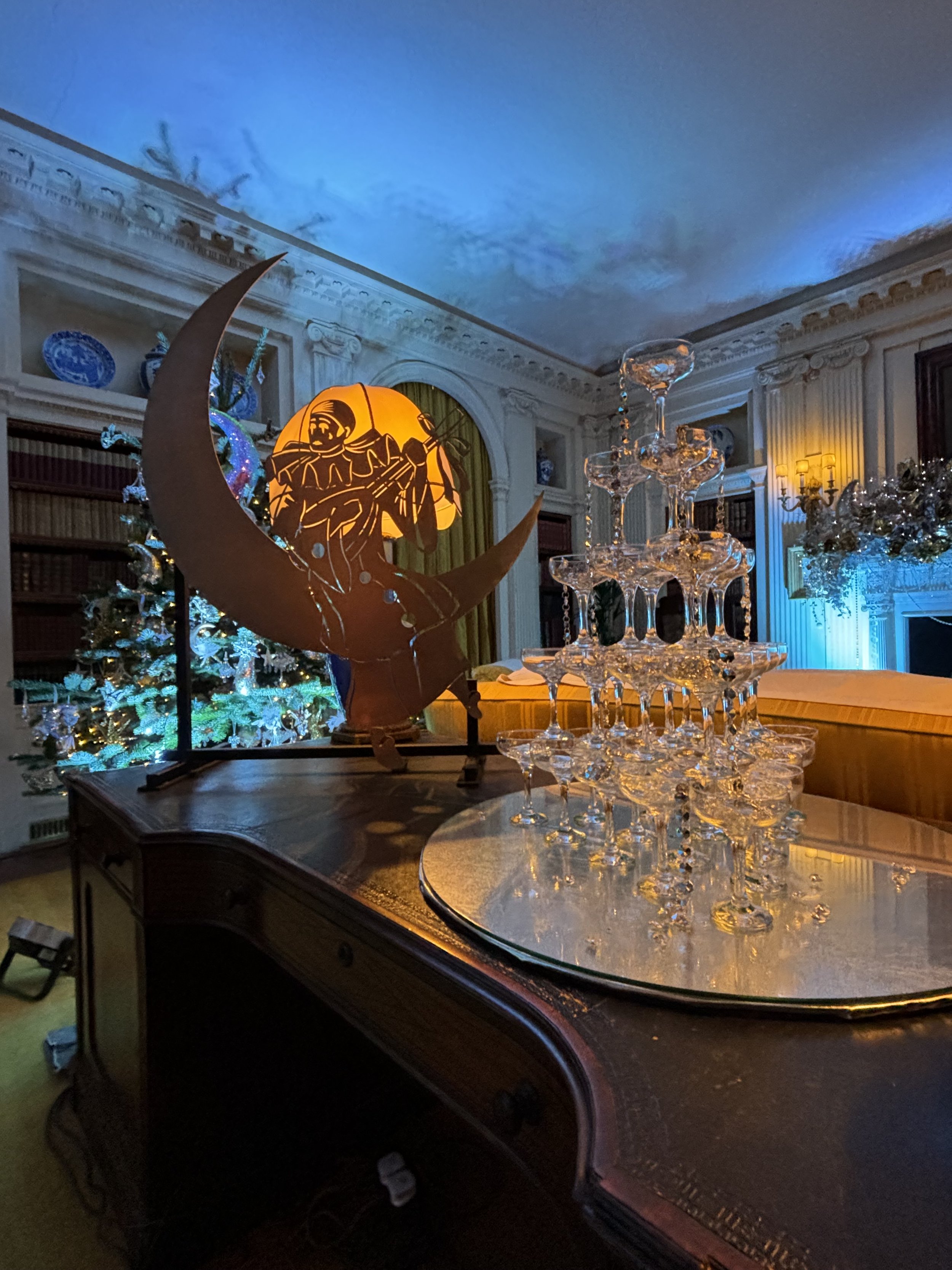 An elegant room with Christmas decorations, including a Christmas tree and a Christmas themed lamp. The room features ornate moldings, bookshelves, and a chandelier, with a champagne tower on a large wooden table.