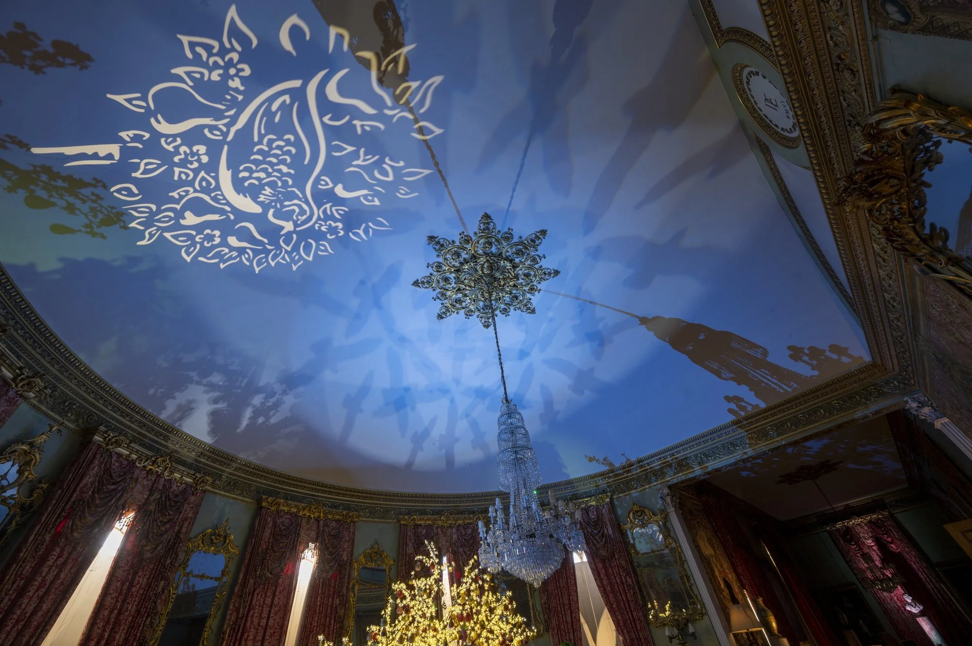 Elegant decorated room with a chandelier, ornate gold moldings, and multiple festive Christmas trees with lights and ornaments. A large ornate clock is visible on the wall.