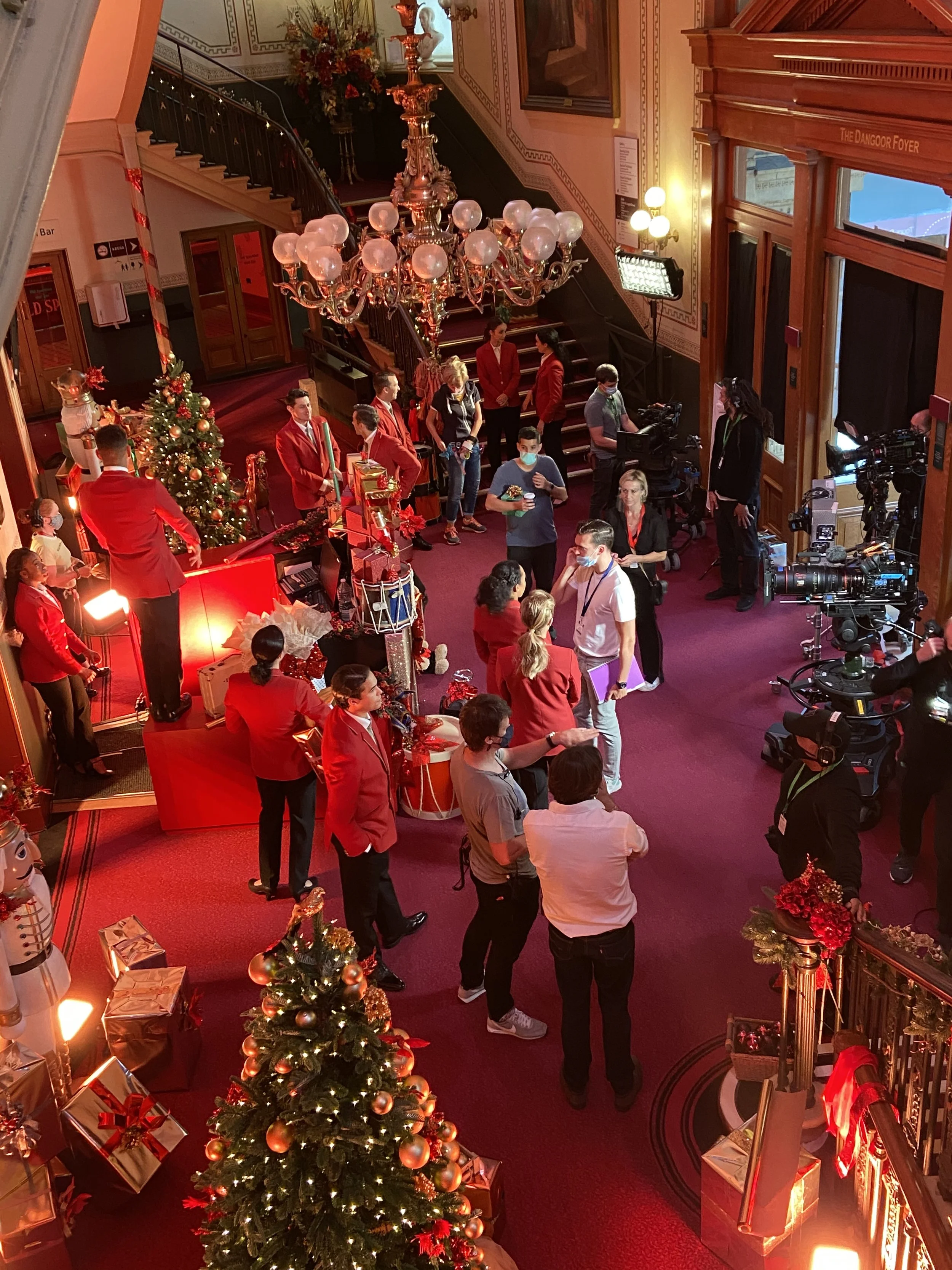 A large, decorated Christmas tree with gold and red ornaments in a festive, elegantly decorated interior. People, many dressed in red, are gathering around, some wearing face masks. There are Christmas presents, a chandelier, and a staircase in the b