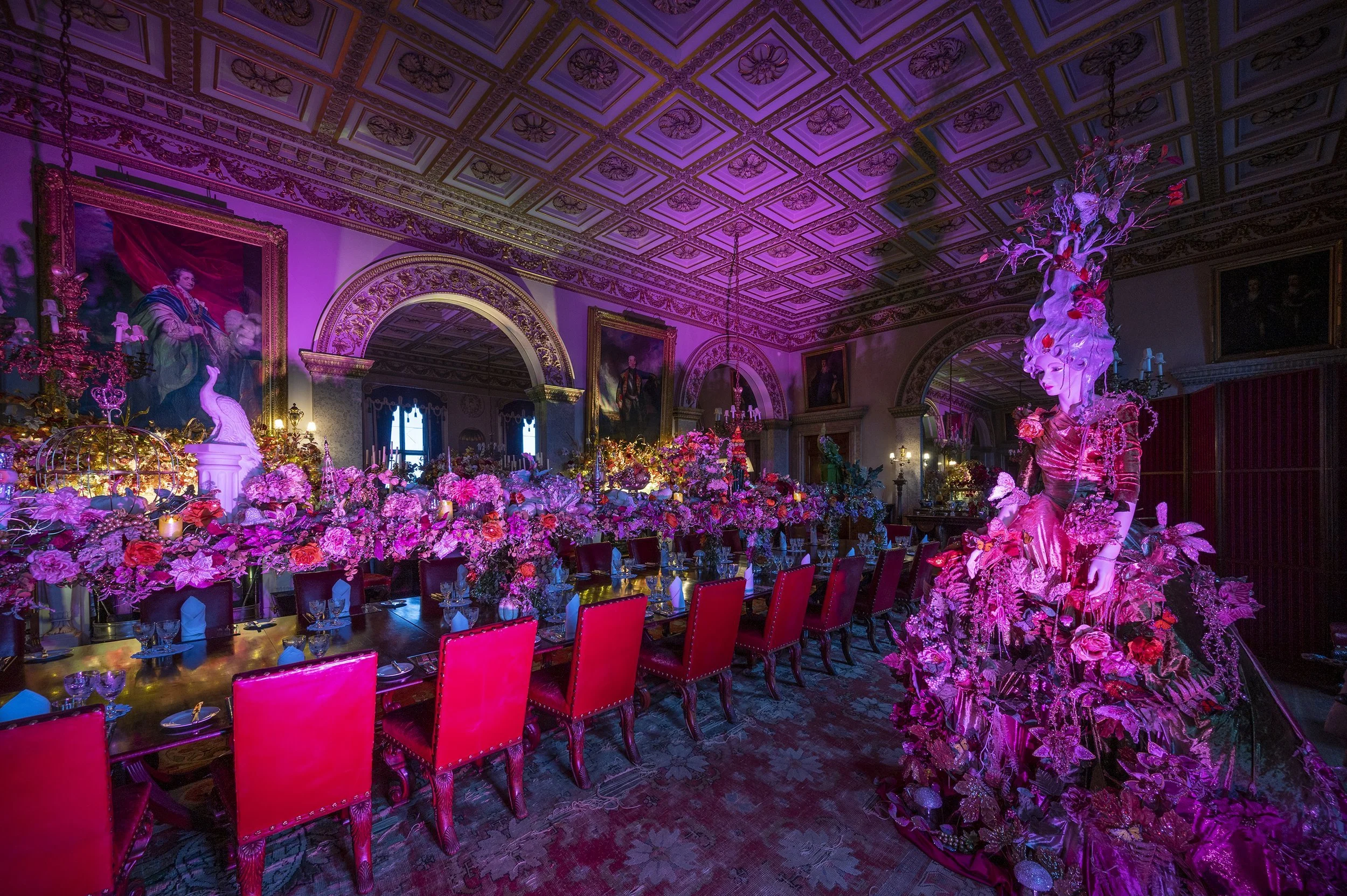 An ornately decorated banquet hall with floral arrangements, a large sculpture of a woman’s face with a floral headdress, and purple lighting.