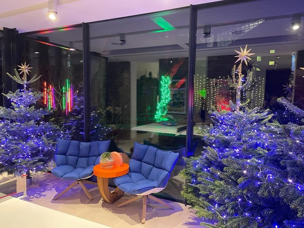 Christmas trees decorated with blue and white lights and star toppers, glass window with colorful neon lights inside a modern indoor space, blue chairs and an orange table with a pineapple-shaped object on top.