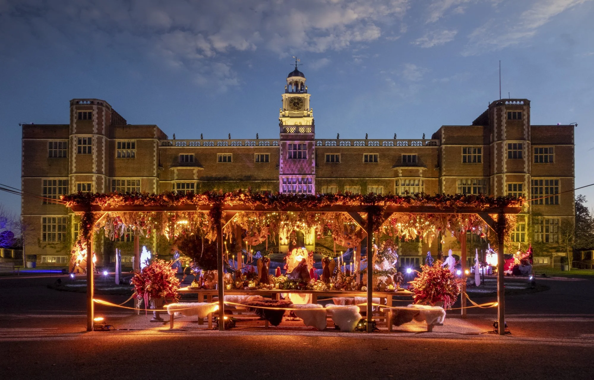 A large historic brick building with a turret and clock tower illuminated at dusk. In front, a festive outdoor display features Christmas nativity scene, decorated with string lights and ornaments, under a wooden structure with greenery and lights.