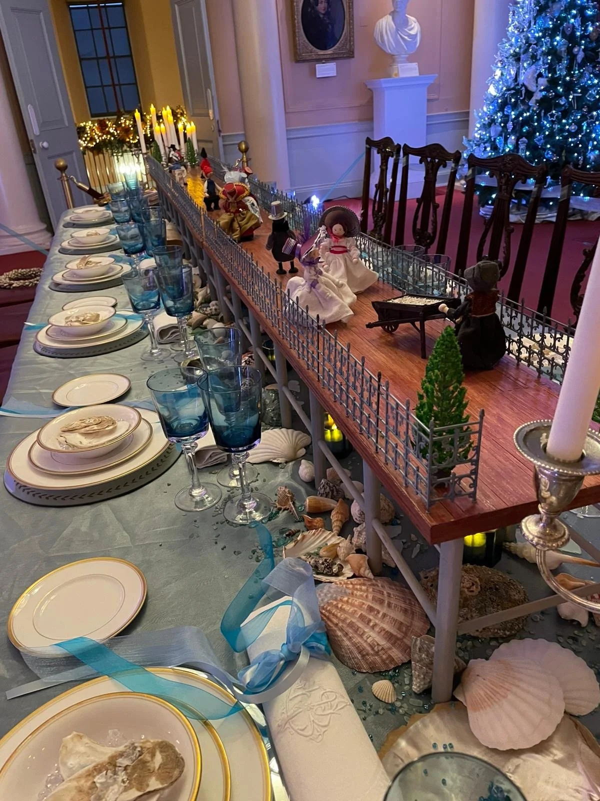 A decorated Christmas table with seashells, blue glasses, and sea-themed decorations, next to a miniature fence and figurines, with a large Christmas tree in the background.