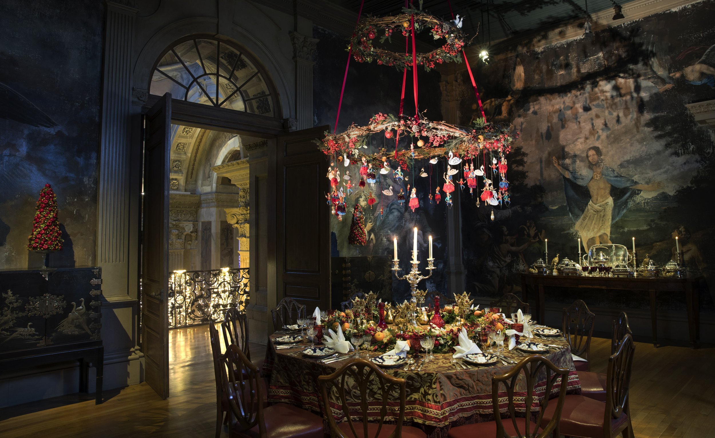 An elegant dinner table setting decorated for Christmas, with a large chandelier adorned with red and gold ornaments and swan decorations hanging from red ribbons. The room has ornate walls, a chandelier, and a mural on the wall, with candles and a C