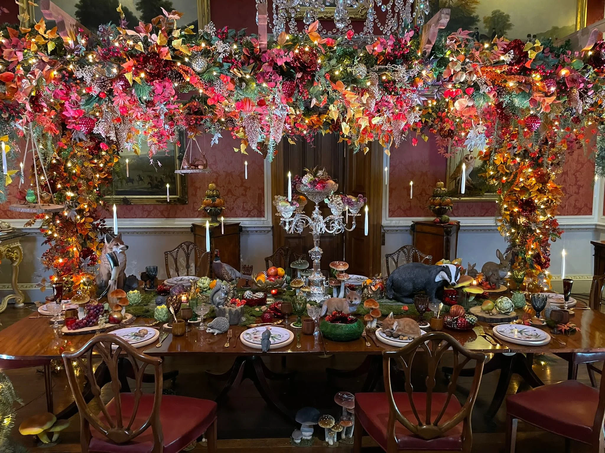 A lavish banquet table decorated with woodland animal figurines, mushrooms, and greenery, with a large floral and light display hanging above in a decorated dining room.
