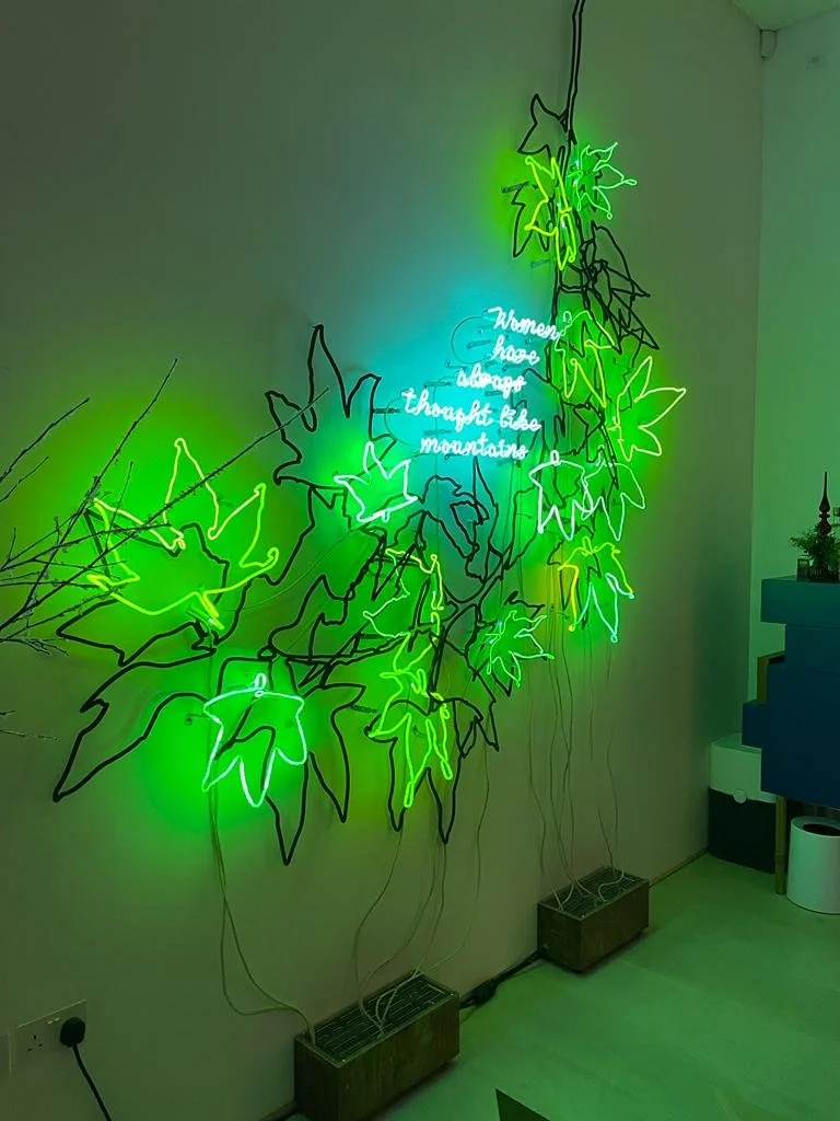 Neon wall art with fluorescent green and blue leaves and a quote that says 'Women have always thought like mountains'.