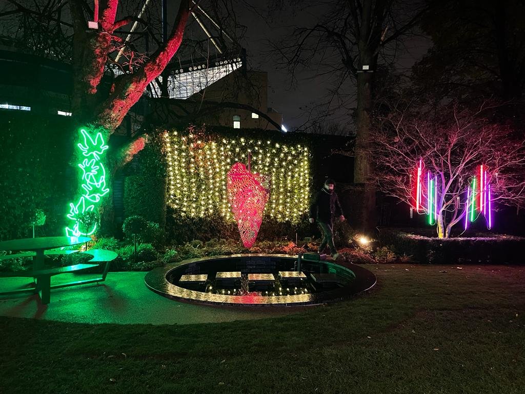 Nighttime garden scene with neon lights, a person walking, decorative lights, and trees illuminated by colorful lights.