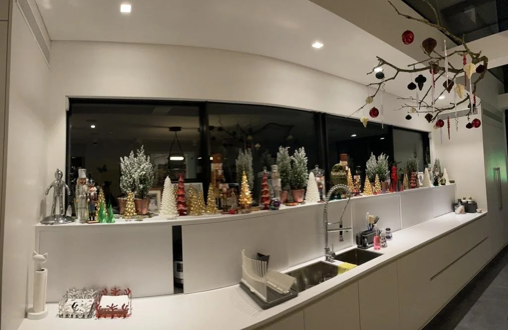 Kitchen window sill decorated with mini Christmas trees, potted plants, and holiday ornaments, with a branch hanging above decorated with small ornaments.