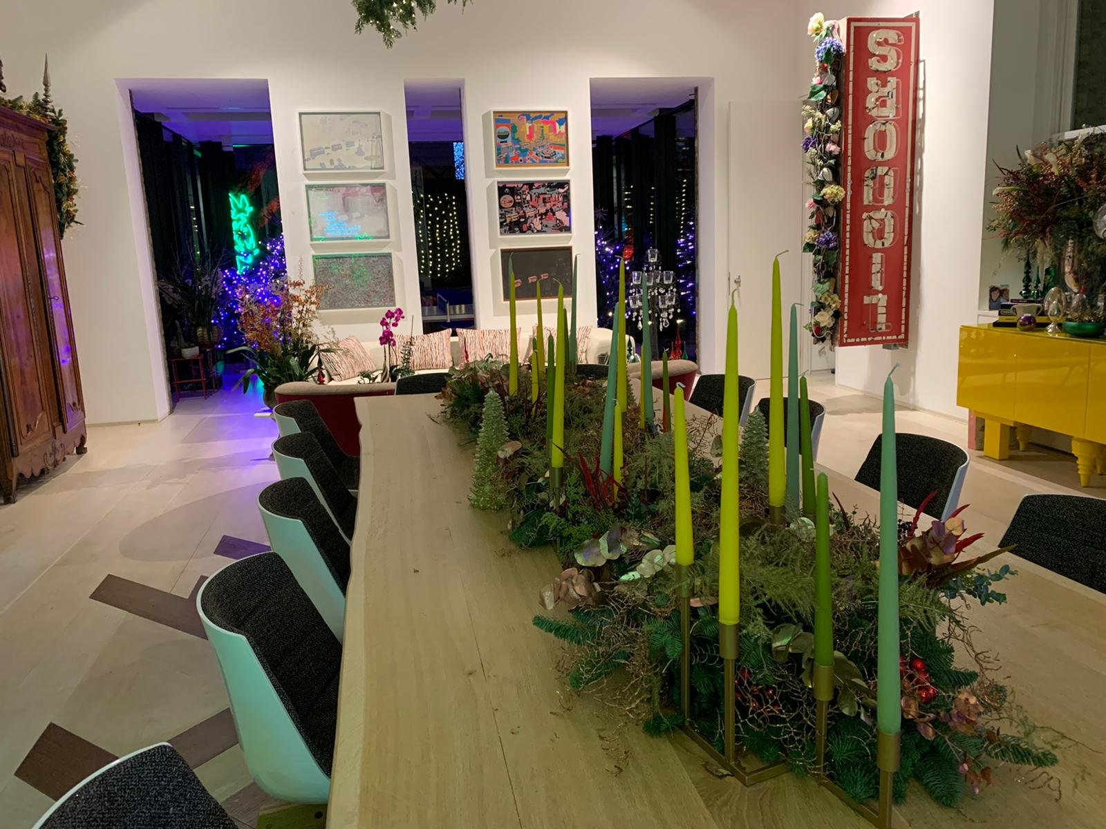 A dining room with a long wooden table decorated with a greenery and candle centerpiece and surrounded by black and white chairs. In the background, there are colorful artworks on the wall, a window with lights outside, and a yellow cabinet on the ri