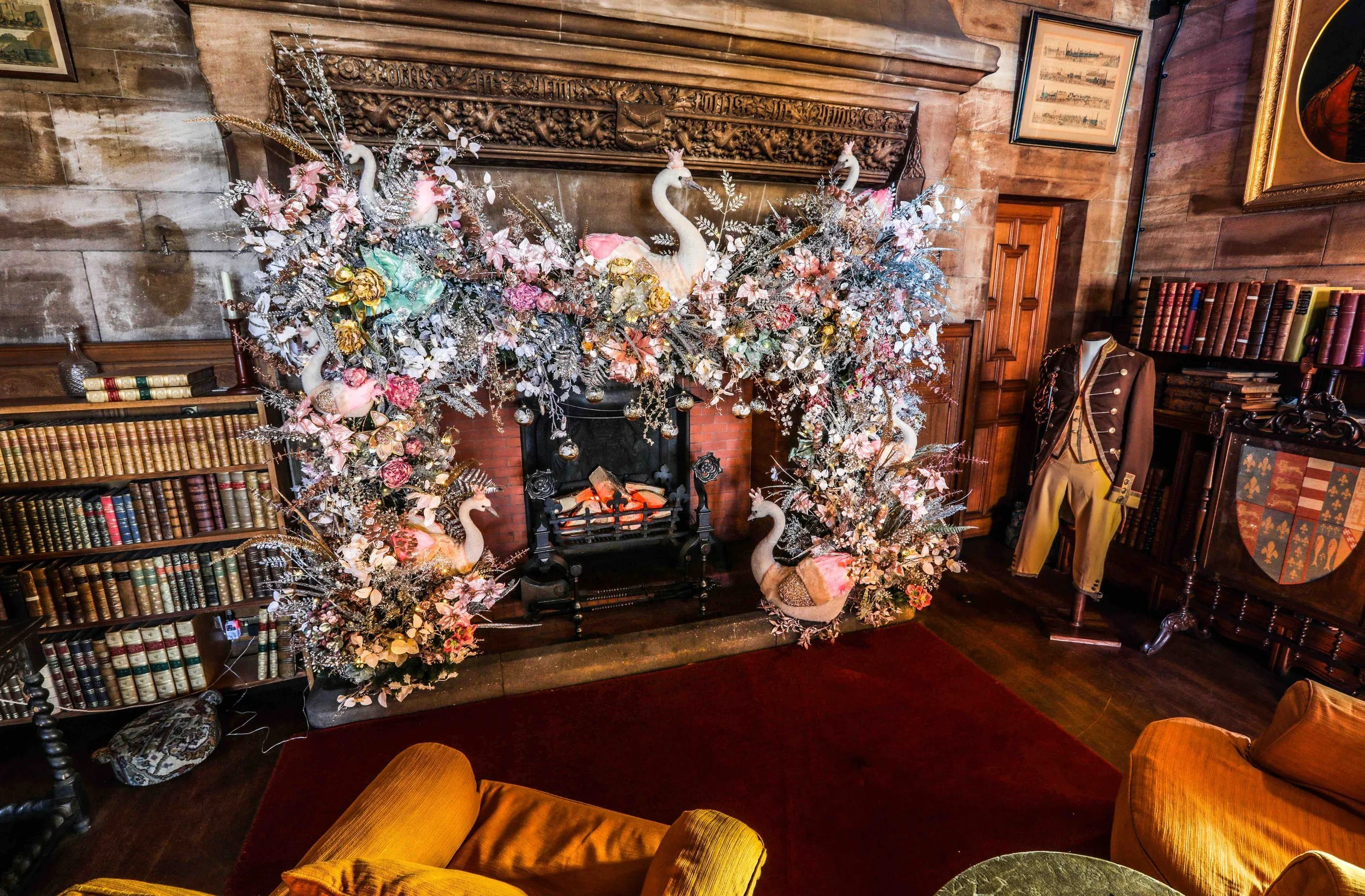An ornate fireplace mantel decorated with an elaborate floral arrangement featuring swan sculptures, pink flowers, gold accents, and silver foliages. The room has wood-paneled walls, framed artwork, a bookshelf with leather-bound books, and antique f