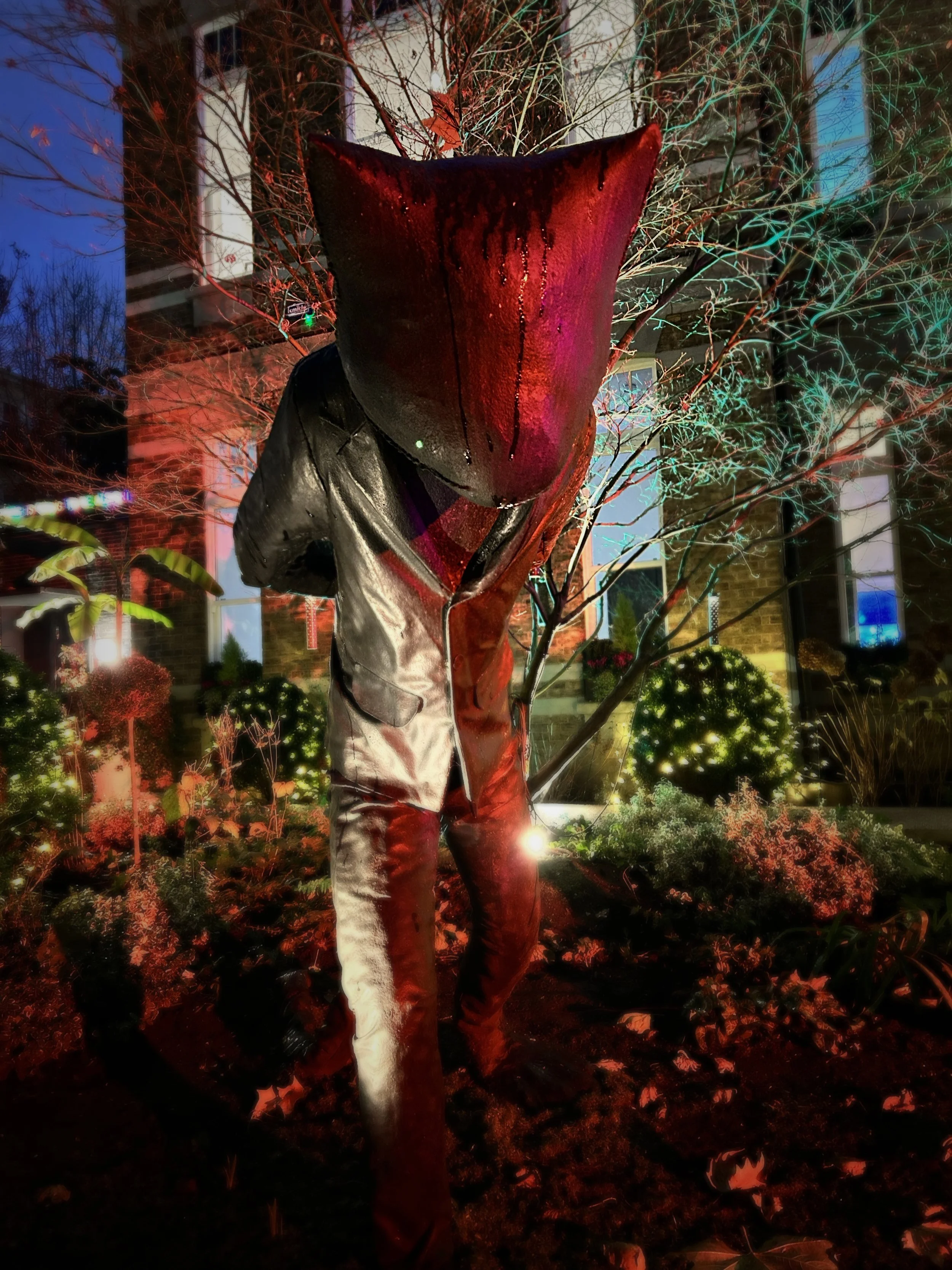 A person standing outdoors at night in front of a house with lit windows, surrounded by trees and bushes illuminated with colorful lights, wearing a black leather jacket and jeans.