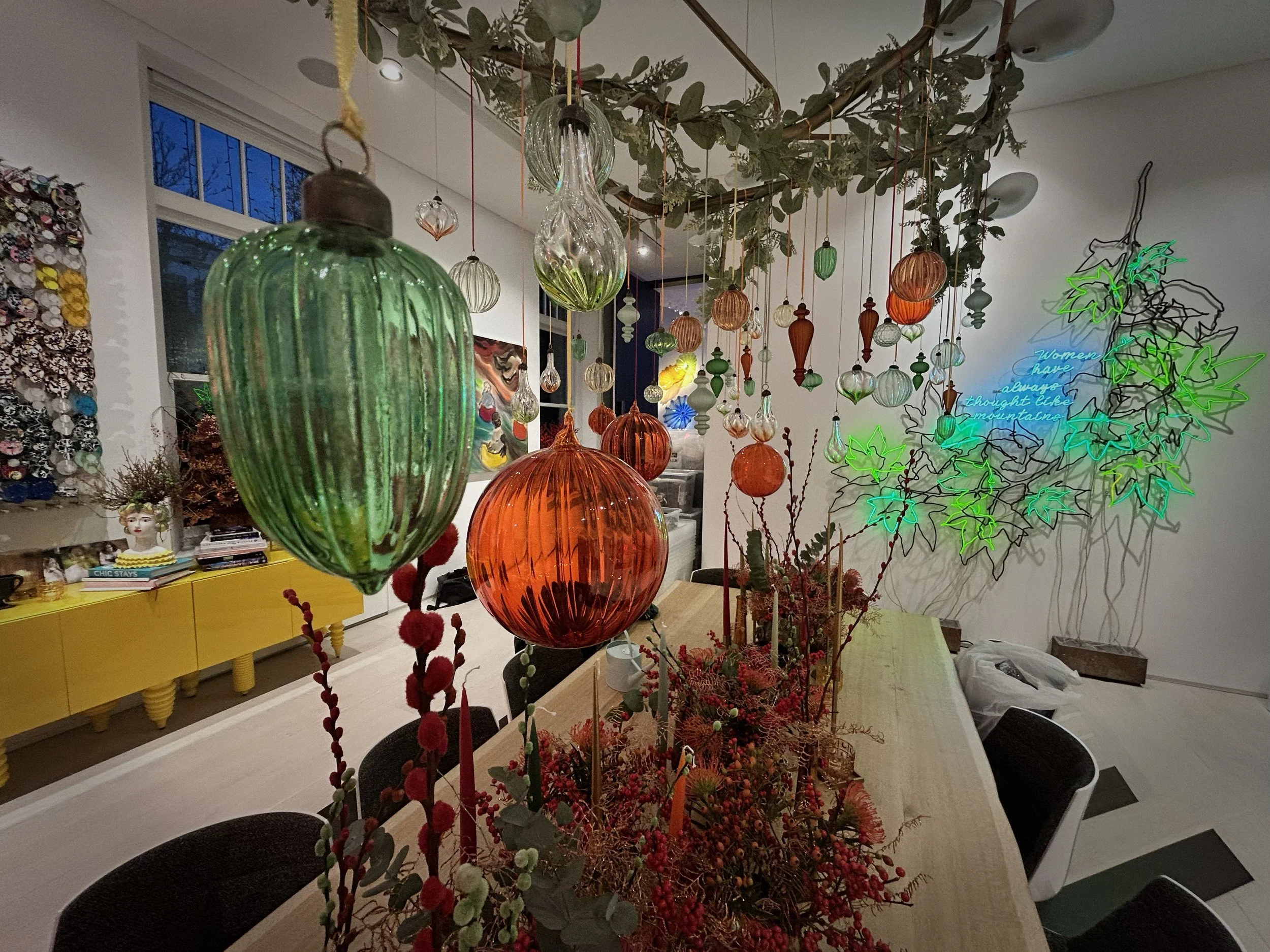 Colorful glass ornaments hanging from a ceiling with green leaves, in a decorated room.