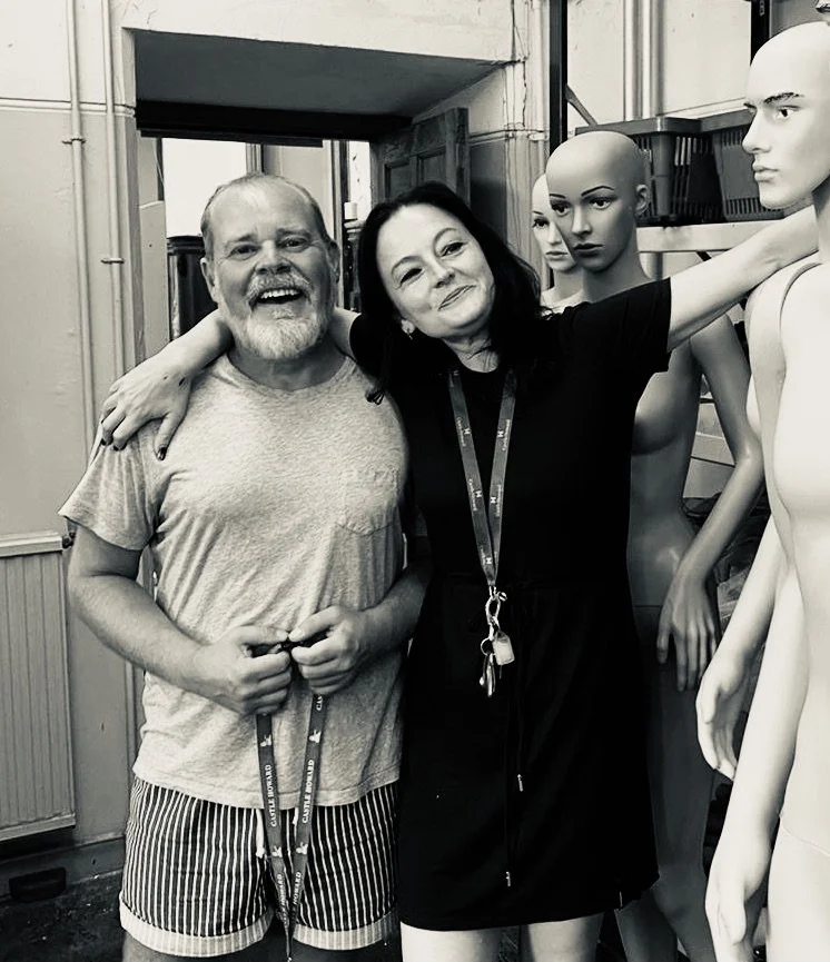 A man and a woman smiling, standing next to each other with their arms around one another. They are standing beside mannequins inside a store or workshop setting.