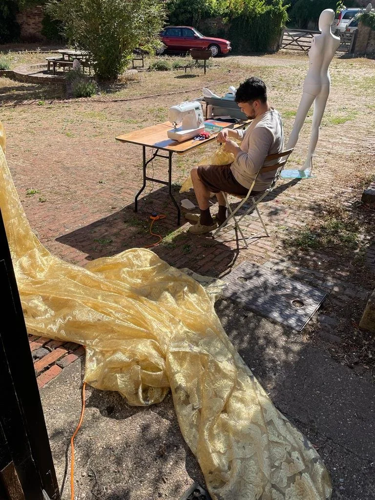 A young man sewing on a sewing machine outdoors at a table, with a yellow fabric draped on the ground nearby. A headless white mannequin is stationed behind him, and a red car and picnic tables are visible in the background.