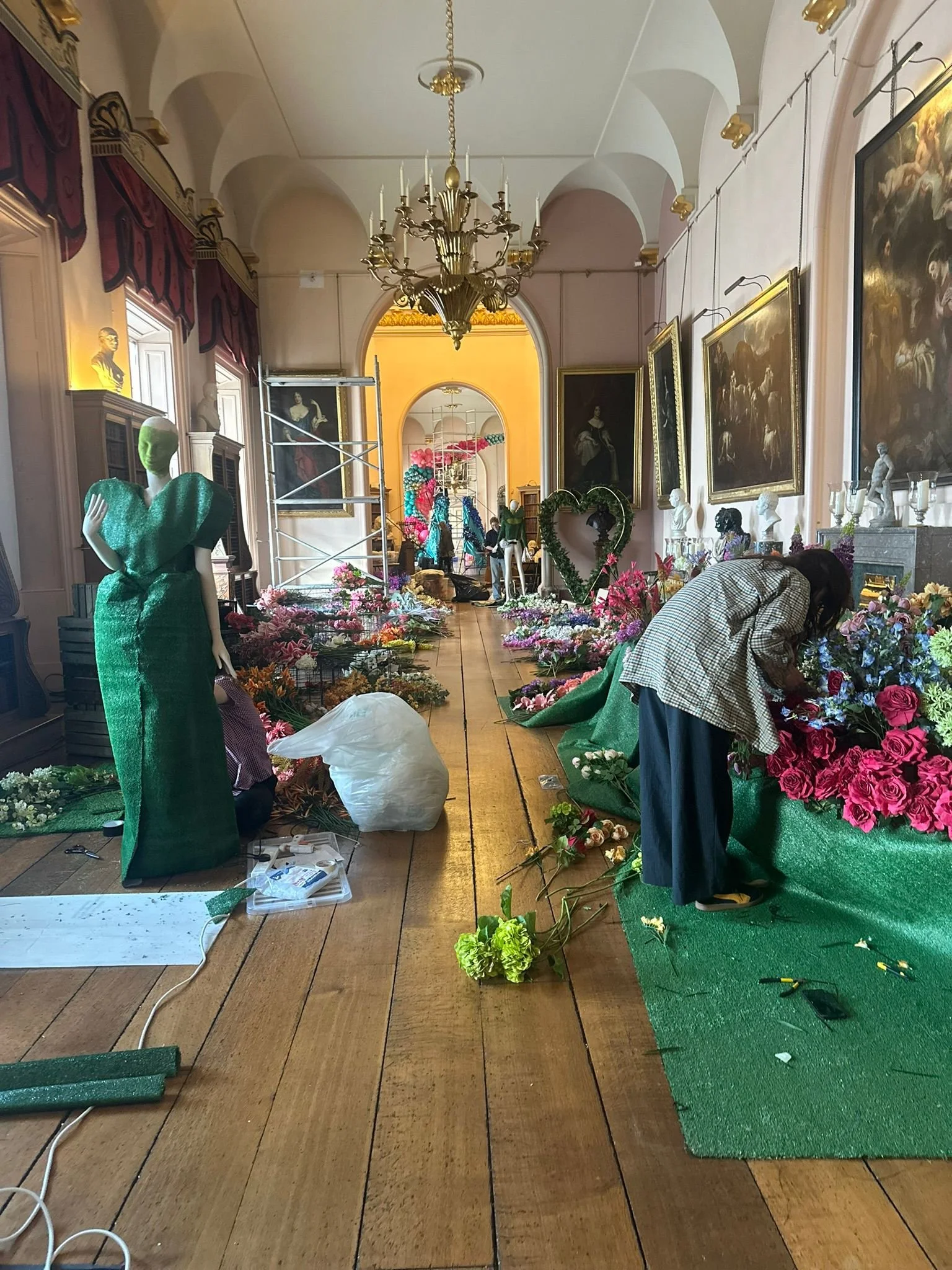 A room decorated with portraits, statues, and floral arrangements in preparation for an event or display. Some people are working on the setup, with tools and materials visible on the floor and tables.
