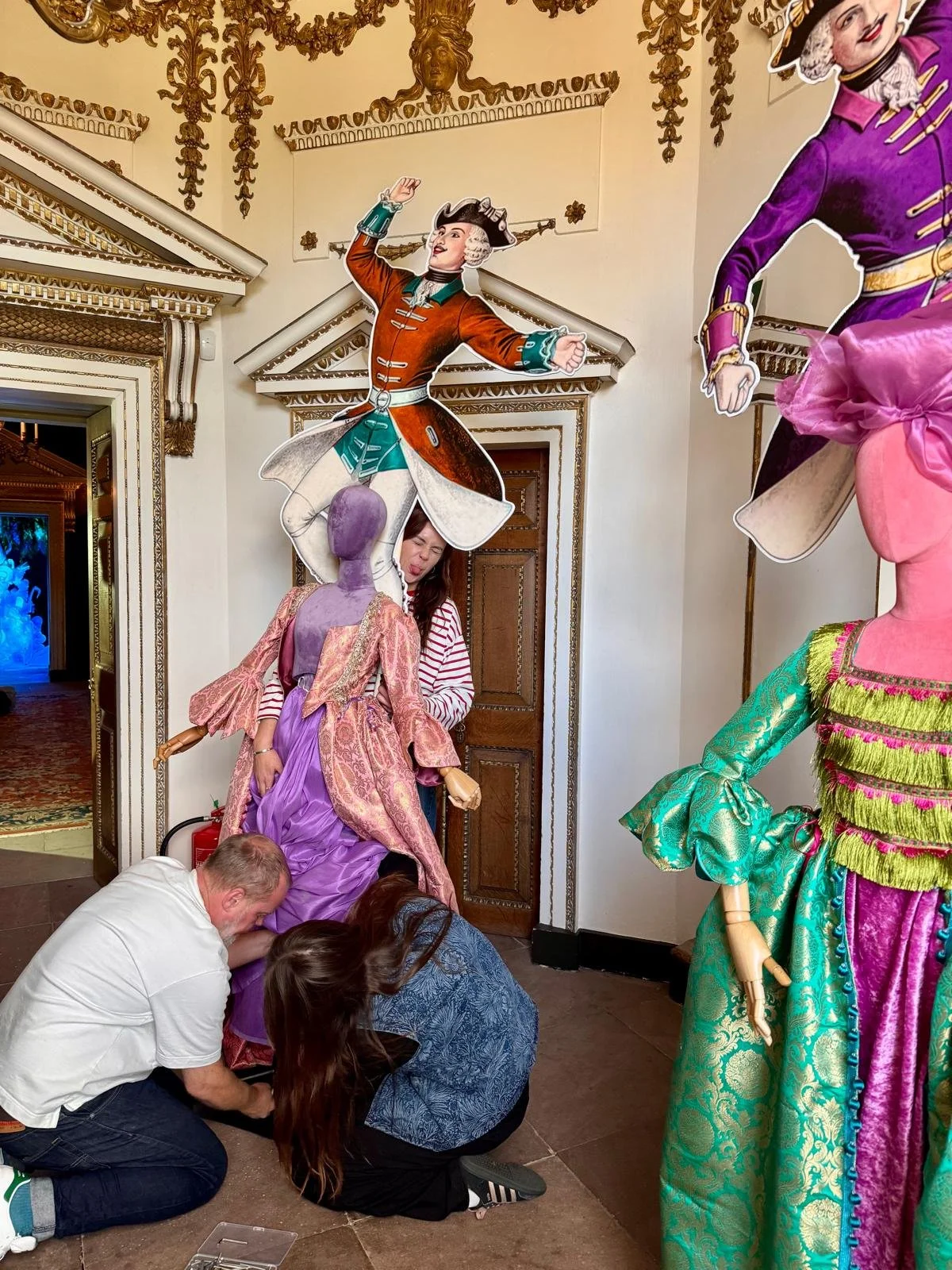 People are installing or repairing a large, ornate puppet or figure dressed in colorful, elaborate costumes, with a woman making a funny face behind it. The scene appears to be inside a decorative, historic building with gold accents and intricate mo