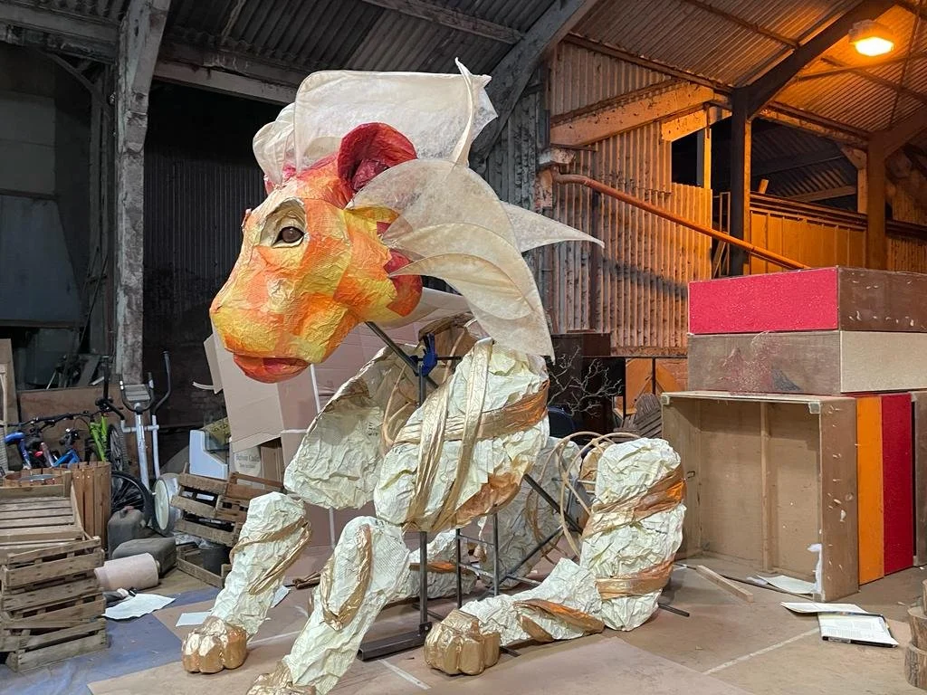 Large papier-mâché lion sculpture in a workshop with wooden pallets, boxes, and equipment behind it.