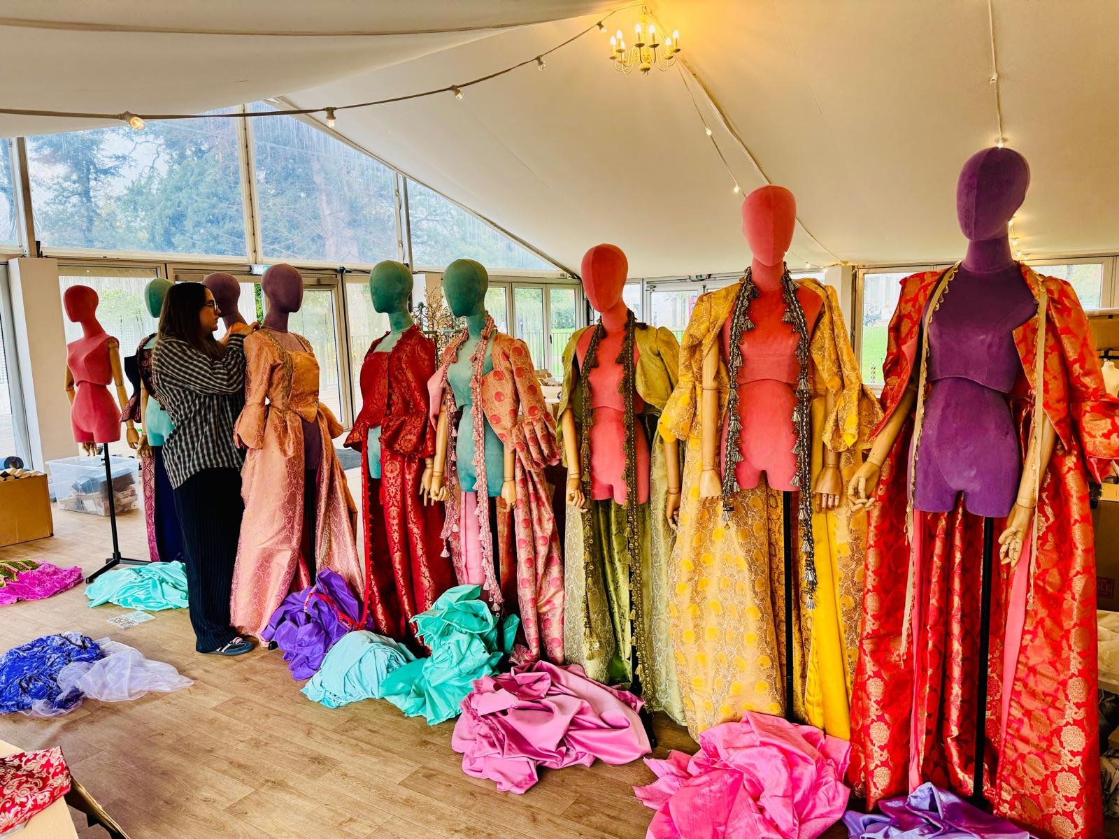 Multiple colorful mannequins dressed in vibrant, patterned dresses and robes are displayed in a room with large windows. A woman is adjusting one of the mannequins. There are piles of fabric on the floor and some stands in the background.
