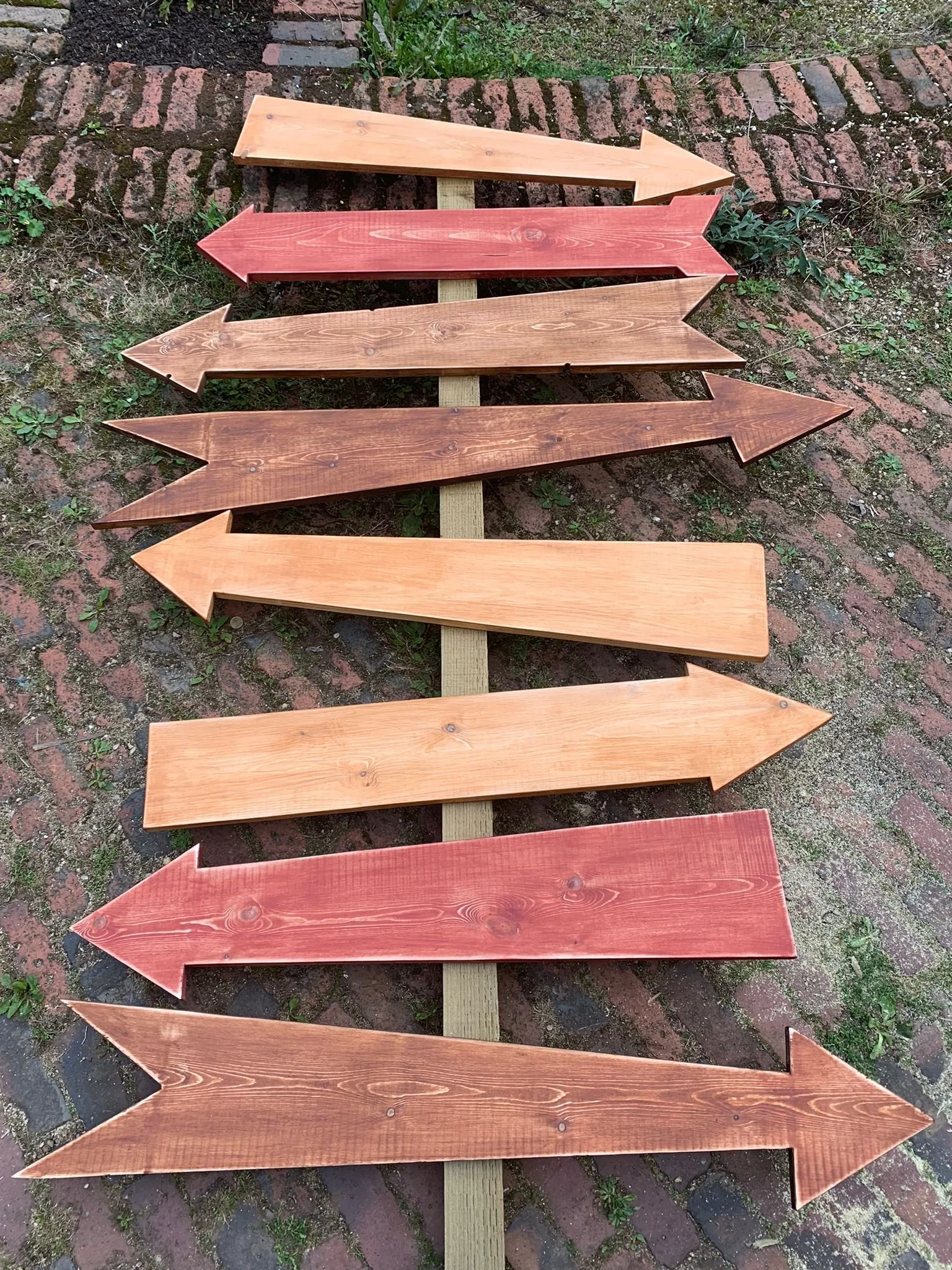 Multiple wooden arrow signs painted in different shades of red and natural wood, arranged in a staggered line on a brick and dirt ground.