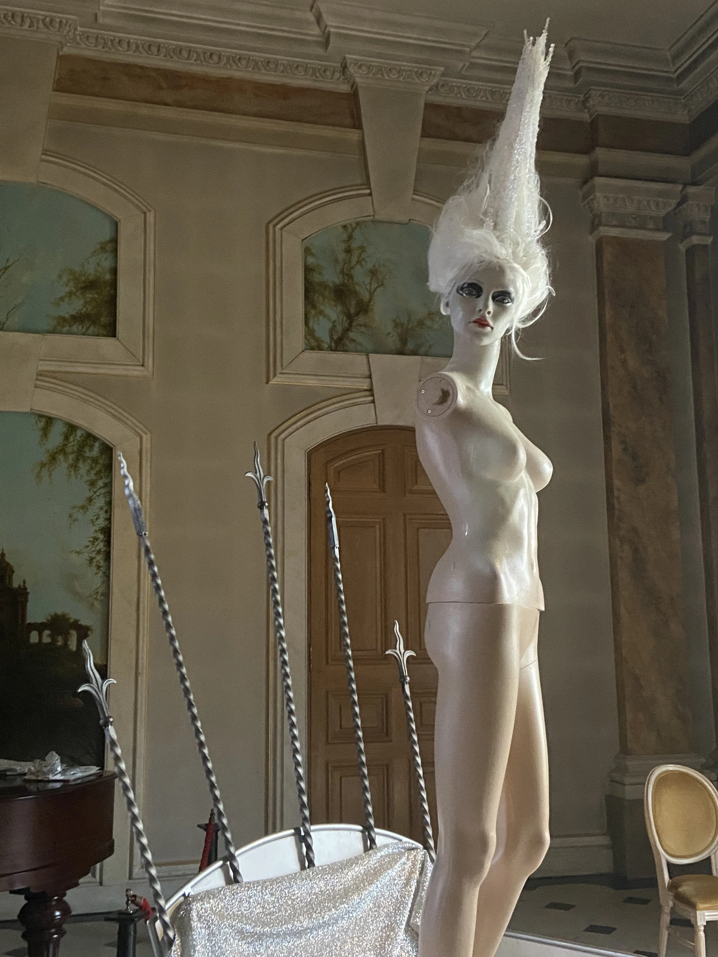 A mannequin with platinum blonde hair styled upwards, standing in a room with ornate crown molding and paintings of trees on the walls.