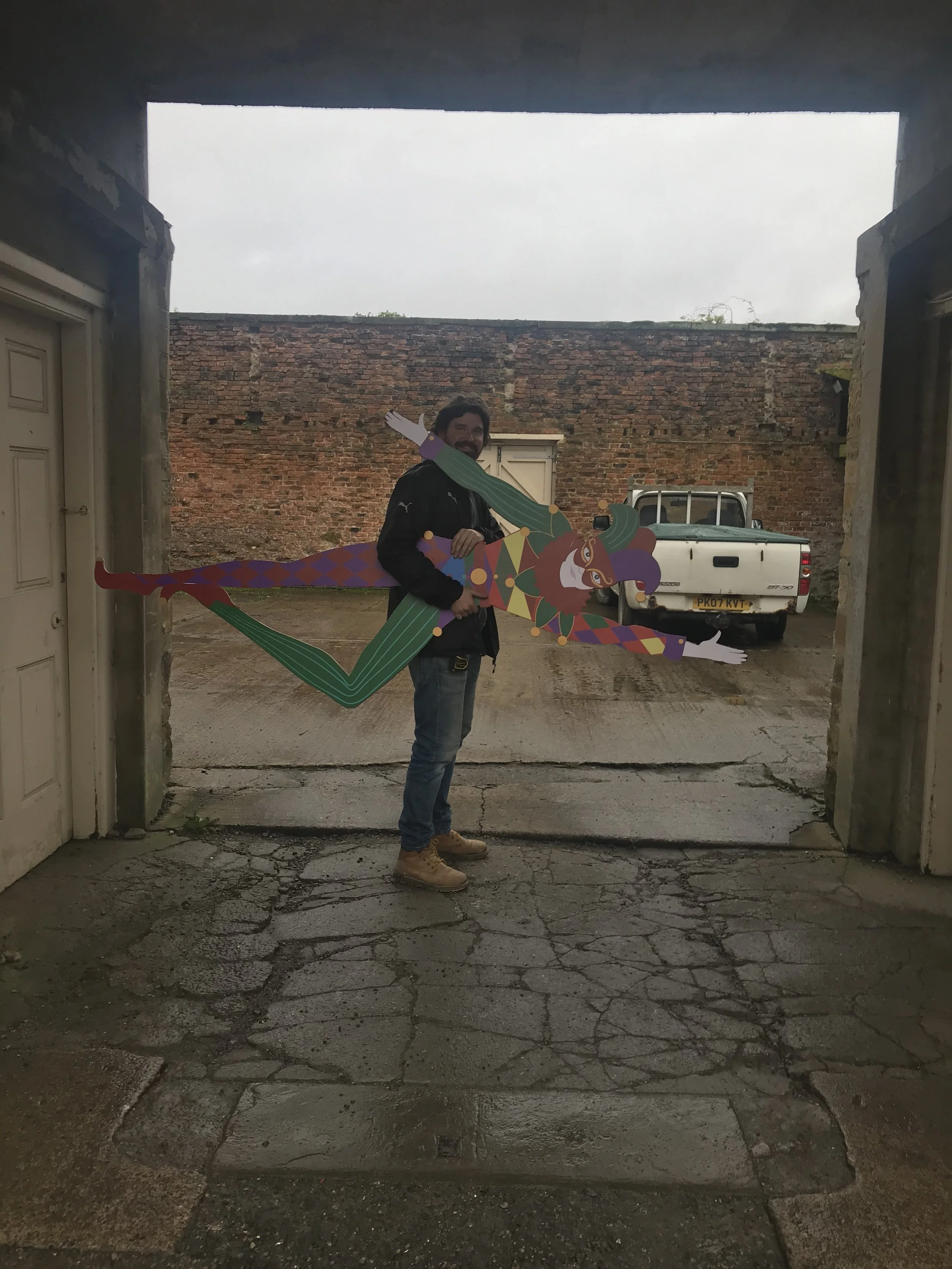 A man holding a colorful, festive cutout of a jester or clown with a pointed hat and a wide smile. The background shows a rustic brick wall, a parked white pickup truck, and the scene is viewed through a large opening in a concrete or stone structure
