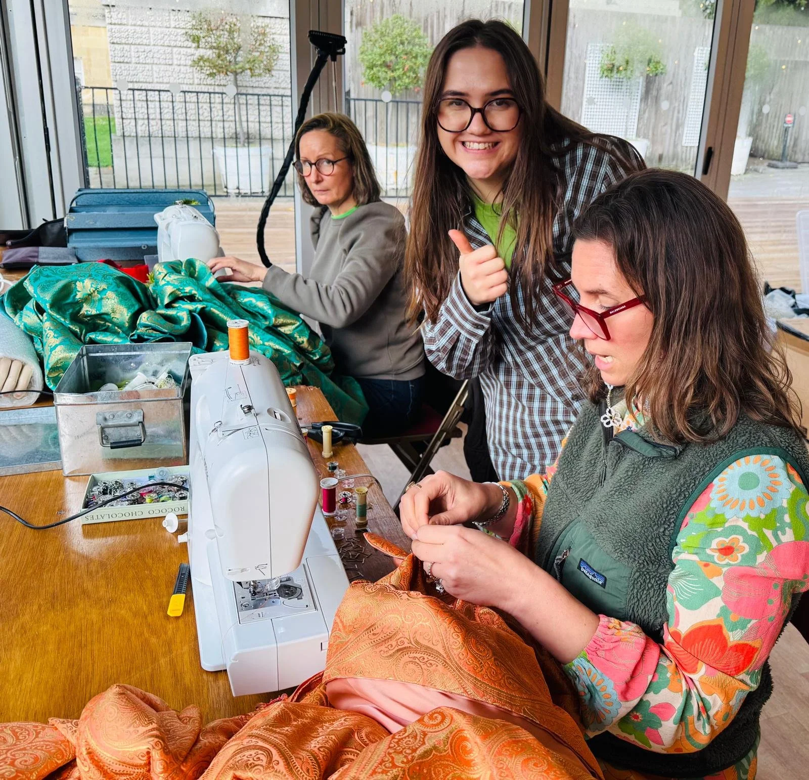 Three women sitting around a table using a sewing machine to work with orange fabric. The woman in the center is smiling and giving a thumbs up, the woman on the right is sewing fabric, and the woman on the left is looking away. The table is filled w