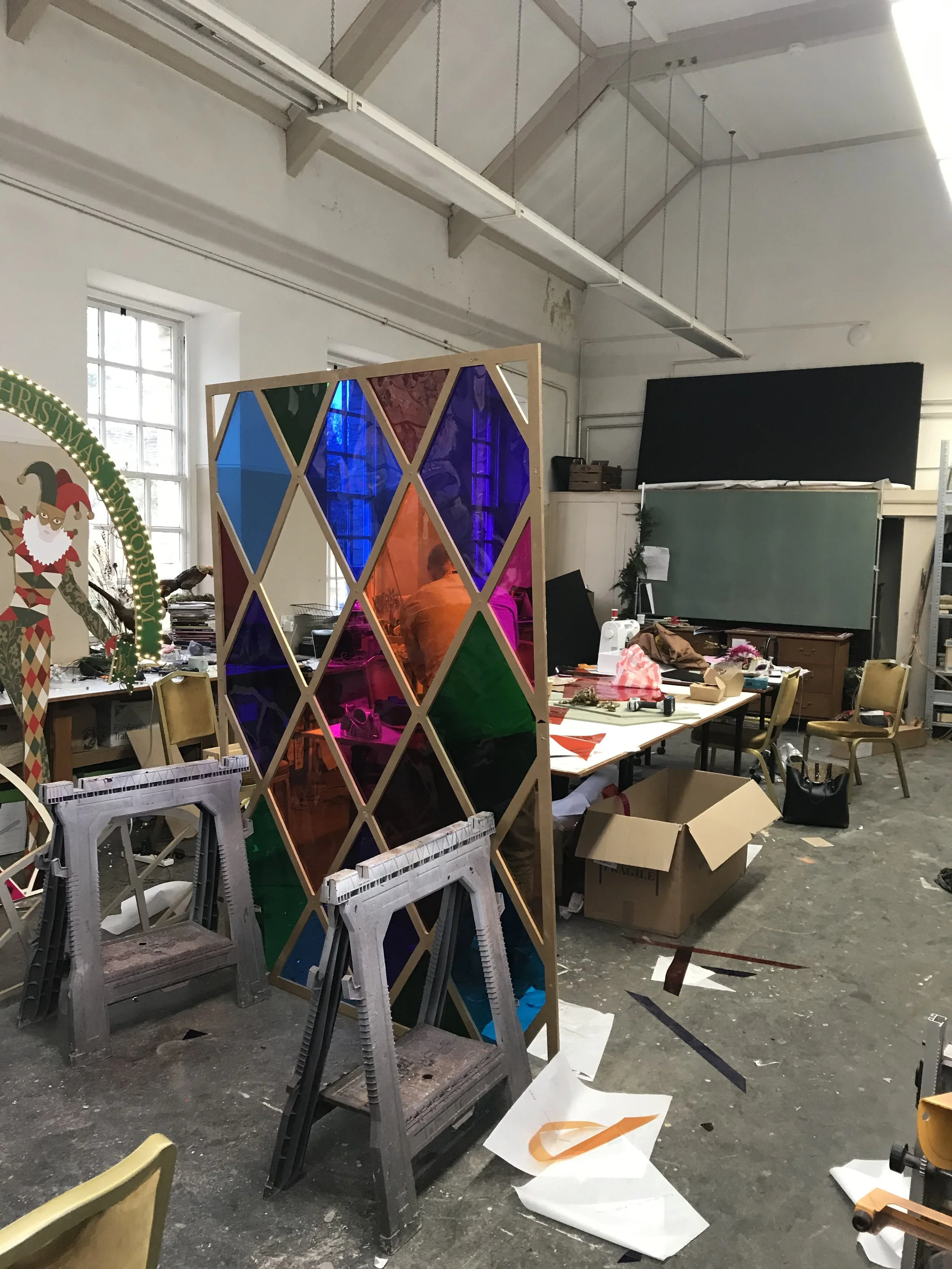 A cluttered workshop or studio with tables, chairs, and colorful stained glass panels. The floor has paper and other materials scattered, and there are shelves and windows in the background.