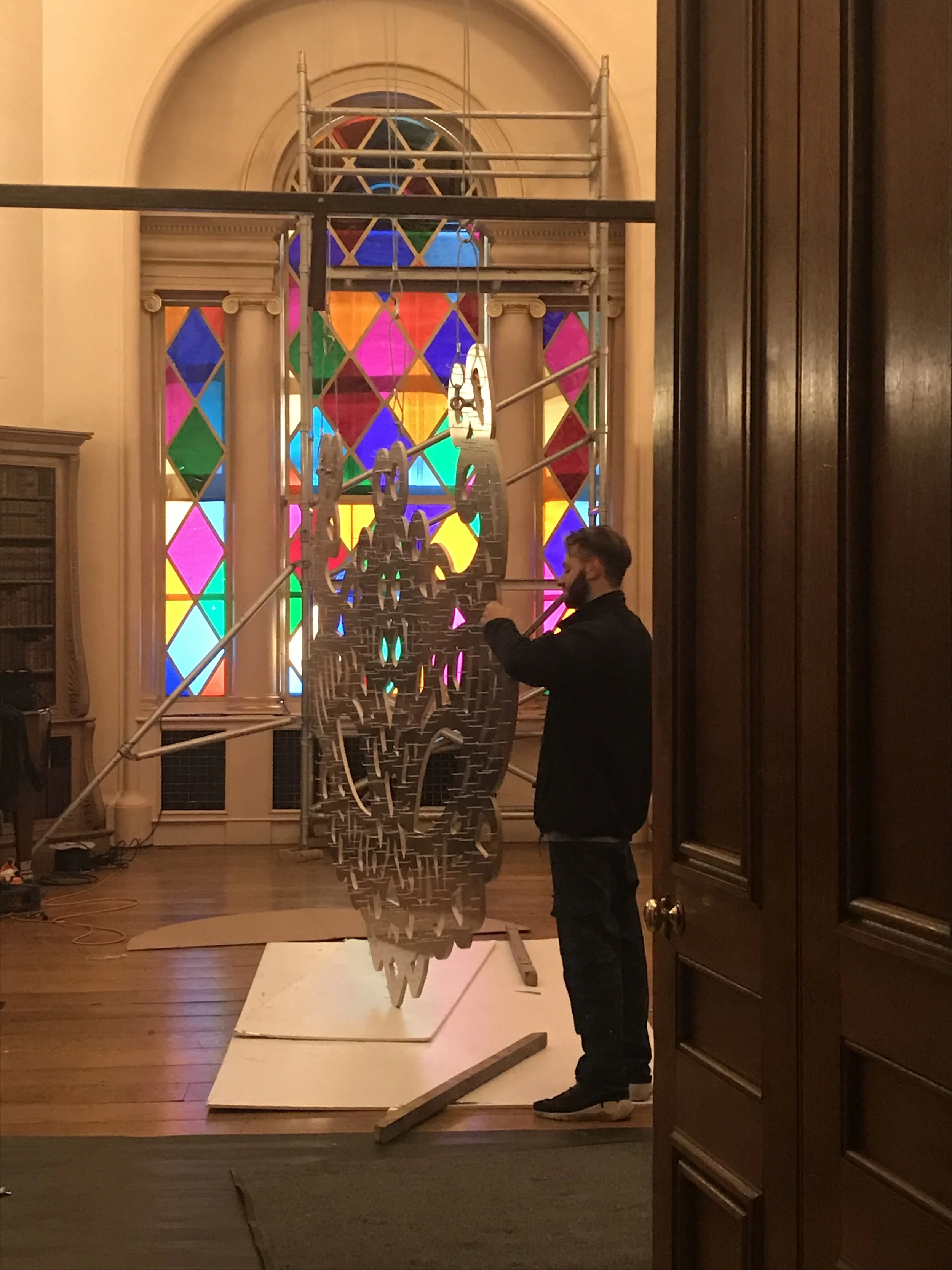 A man with a beard and dark clothing standing inside a building with wooden objects, holding a metal rod, with a colorful stained glass window in the background.