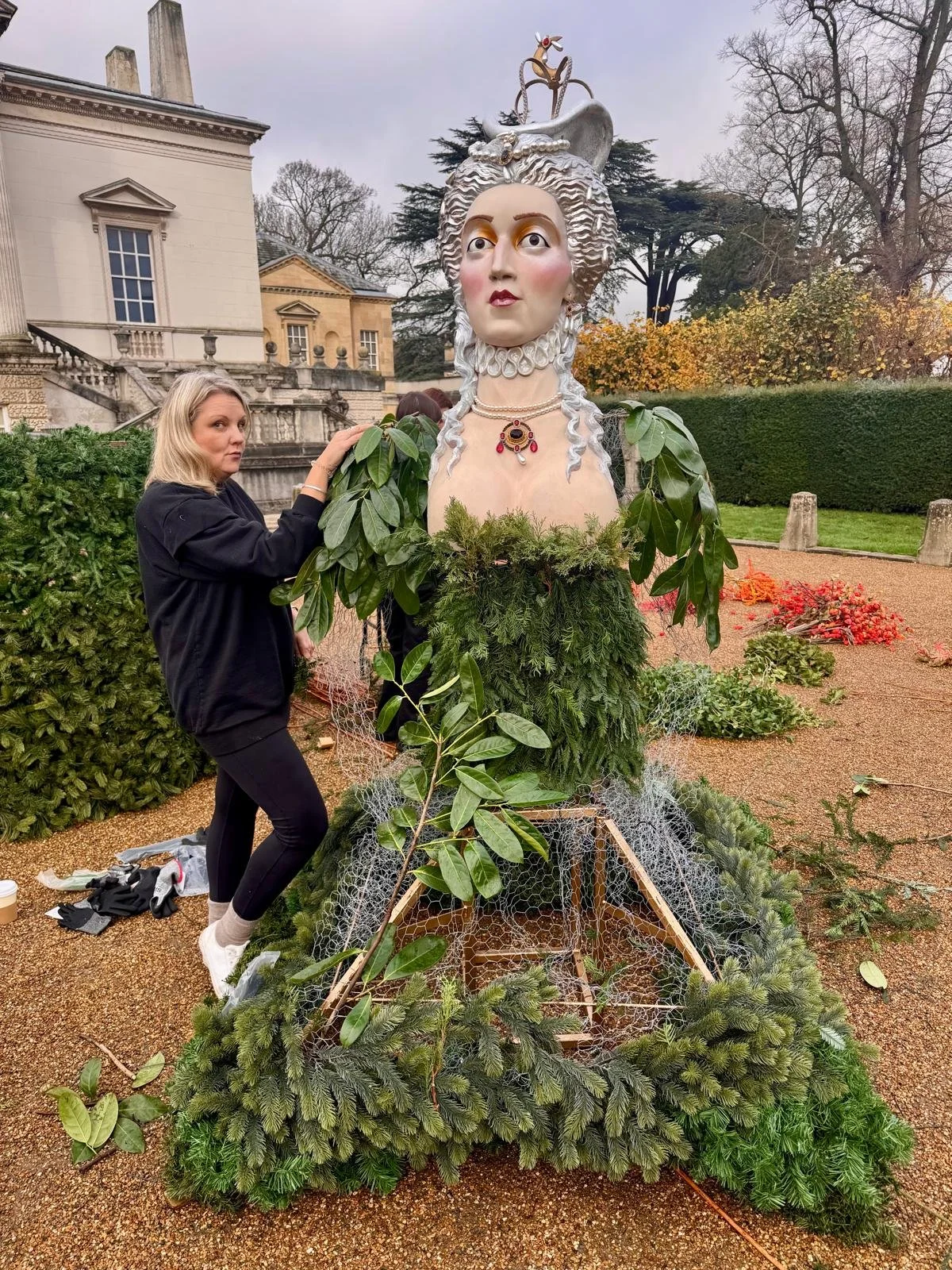 A woman is working on a large holiday decoration of a female figure with elaborate makeup, jewelry, and a crown, surrounded by greenery and ornaments outdoors during the daytime.