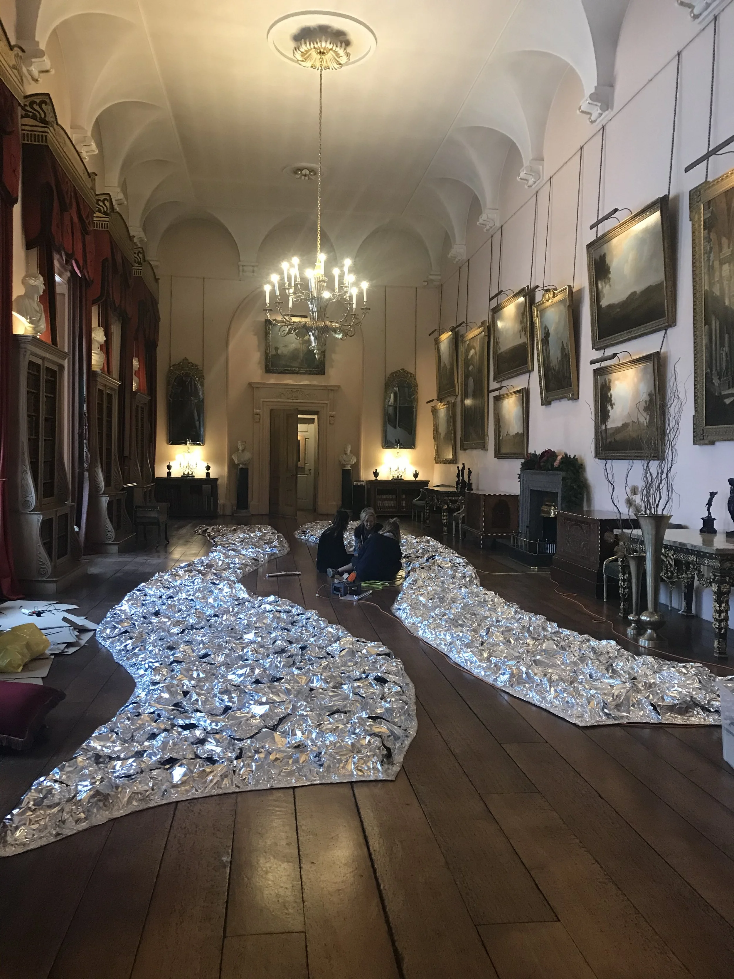 A large, elegant room with a high, arched ceiling, chandelier, and framed paintings on the wall. Two people are sitting on the wooden floor surrounded by crumpled aluminum foil, with some scattered papers and objects nearby.