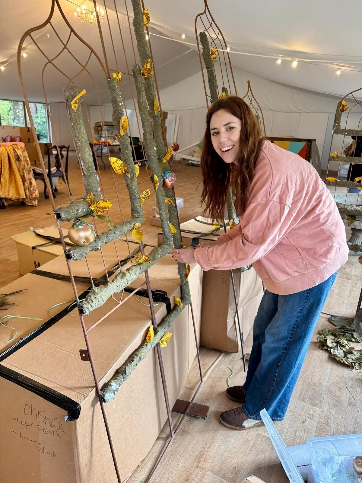 A woman with long brown hair and a pink jacket is working on a tree branch sculpture decorated with yellow butterflies and ornaments inside a large white tent. The sculpture is supported by a metal frame, and the woman is smiling at the camera while 