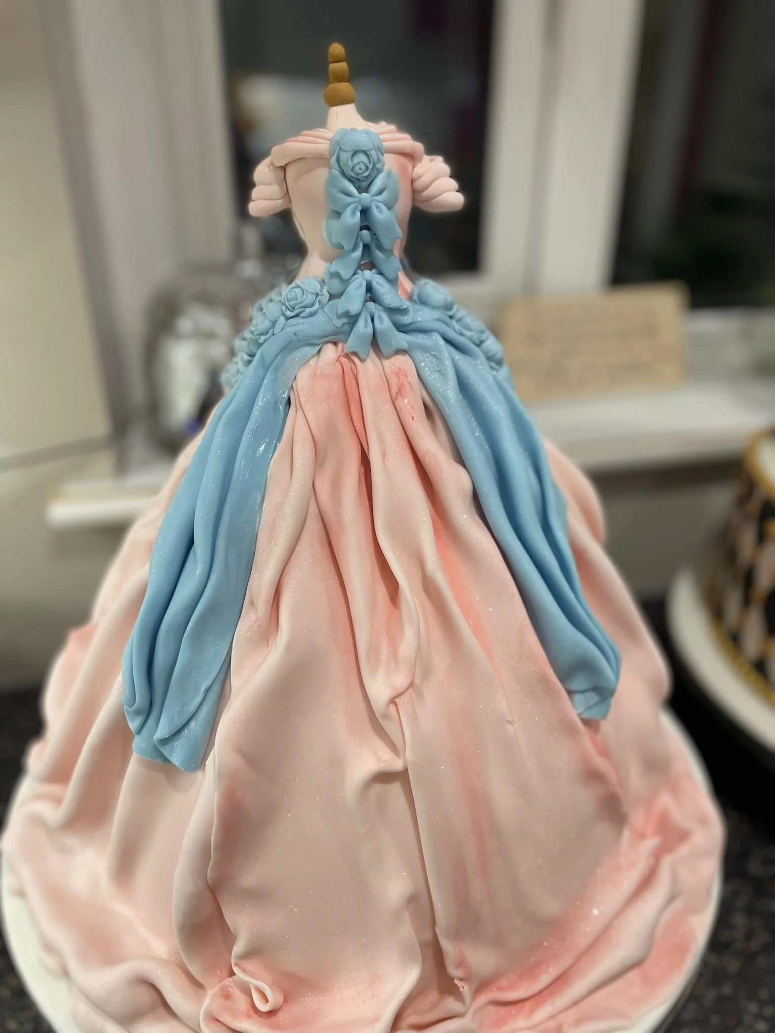Close-up of a cake decorated to look like a gown with pink, blue, and cream-colored draped icing, topped with a unicorn horn and roses.