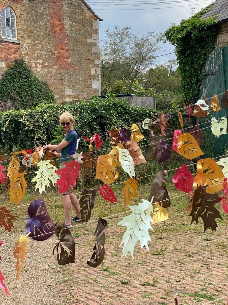 A person with short blonde hair wearing sunglasses, a blue shirt, and shorts, standing outdoors behind colorful paper leaf decorations hanging on strings. The background includes a stone and brick building, greenery, and trees.