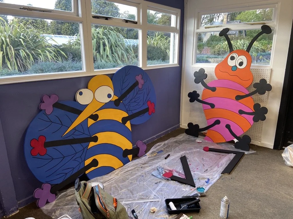 Two large colorful cartoon bee and butterfly cutouts with exaggerated eyes and smiley faces, decorated with flowers, placed against a wall near windows, on a plastic-covered floor with painting supplies nearby.