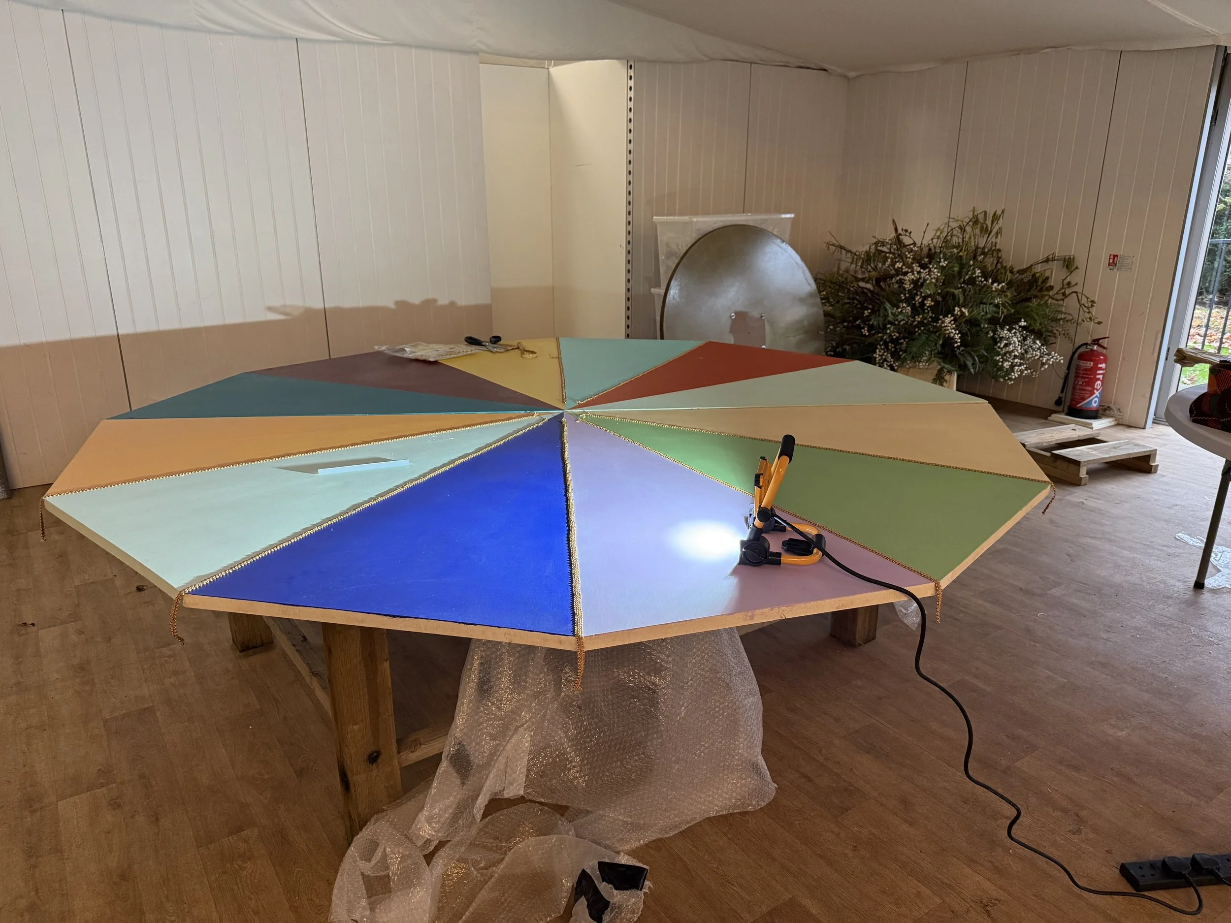 A large round table with a colorful segmented surface, featuring sections in blue, green, yellow, orange, and purple. On the table, there are scissors, a yellow item, and a black marker. In the background, there is a flower arrangement with greenery 