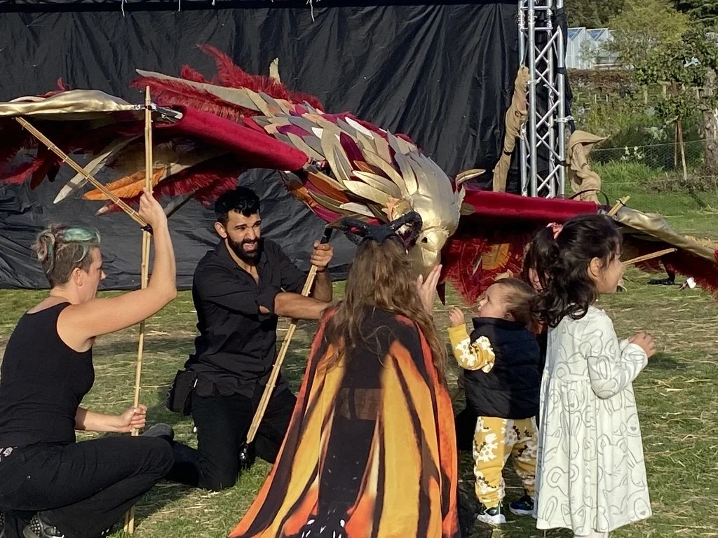 Children and adults participate in a performance with a large, colorful dragon puppet outdoors on grass. The puppeteers are controlling the dragon with poles, and some children are dressed in costumes.